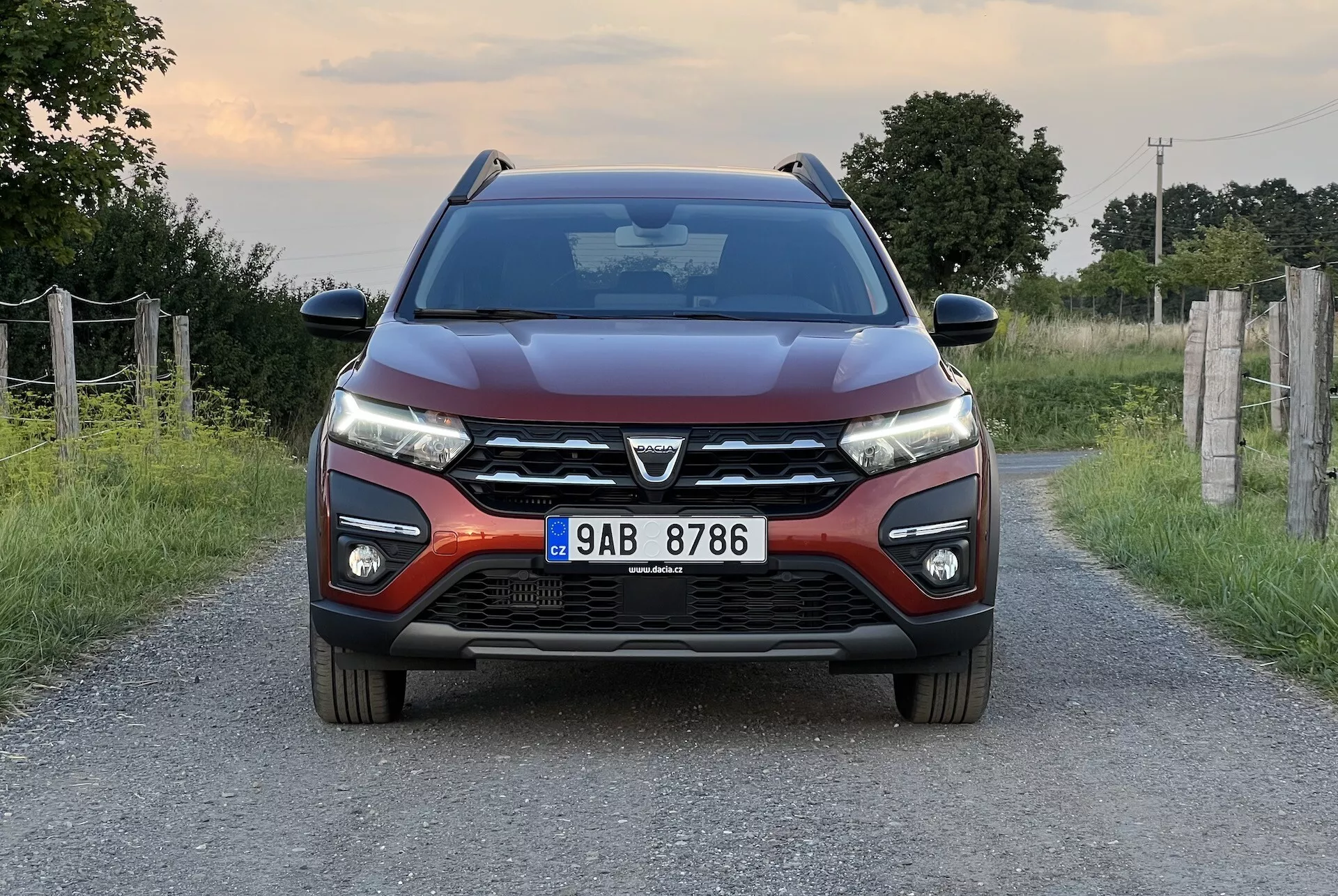 Dacia Jogger 2022 posizionata su una stretta strada rurale, mostrando una vista frontale simmetrica che evidenzia il design moderno dell'auto, incluse una griglia anteriore prominente con un grande emblema, fari LED eleganti, linee sagomate sul cofano, paraurti anteriore con luci fendinebbia e sensori di parcheggio, e specchietti laterali con indicatori di direzione integrati.
