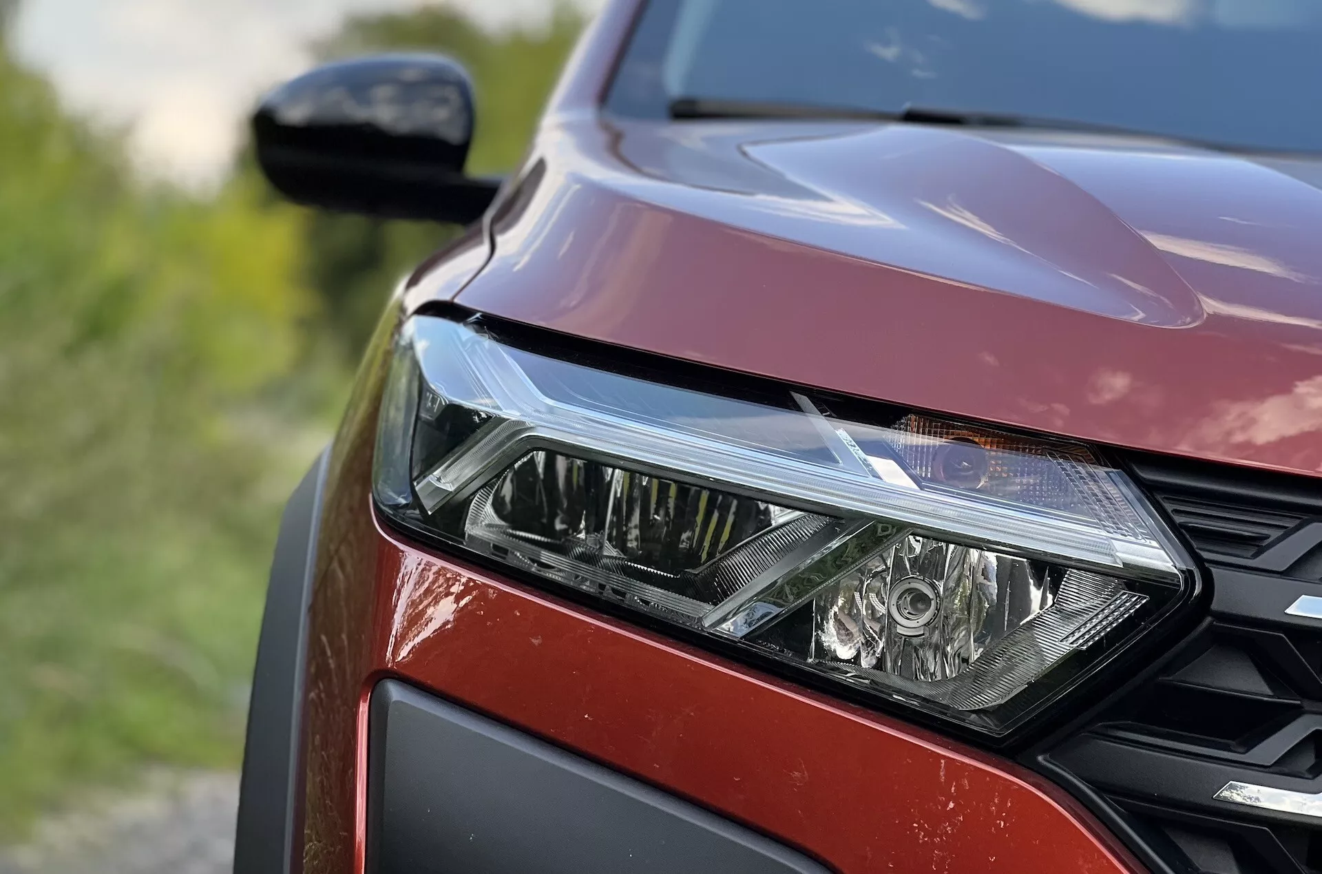 Vista ravvicinata del lato anteriore sinistro della Dacia Jogger 2022 evidenziando il gruppo ottico LED, il motivo a griglia a nido d'ape e la finitura della vernice rossa metallizzata