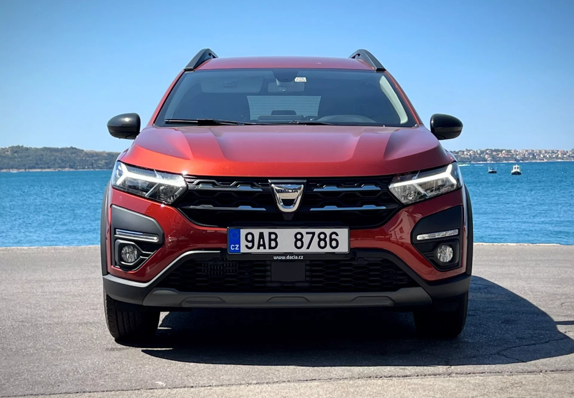 Dacia Jogger 2022 che mette in mostra il suo design moderno da crossover e le avanzate caratteristiche tecnologiche in un tranquillo scenario acquatico, con un griglia prominente con logo, fari LED, luci di posizione, potenziali sensori ADAS, prese d'aria per il raffreddamento e il radar, schema di colori bicolore rosso e nero, cofano aerodinamico, equipaggiato con tergicristalli che indicano una possibile tecnologia sensibile alla pioggia, specchietti laterali con indicatori di direzione integrati potenzialmente contenenti tecnologia di monitoraggio dell'angolo cieco, sotto cieli sereni.