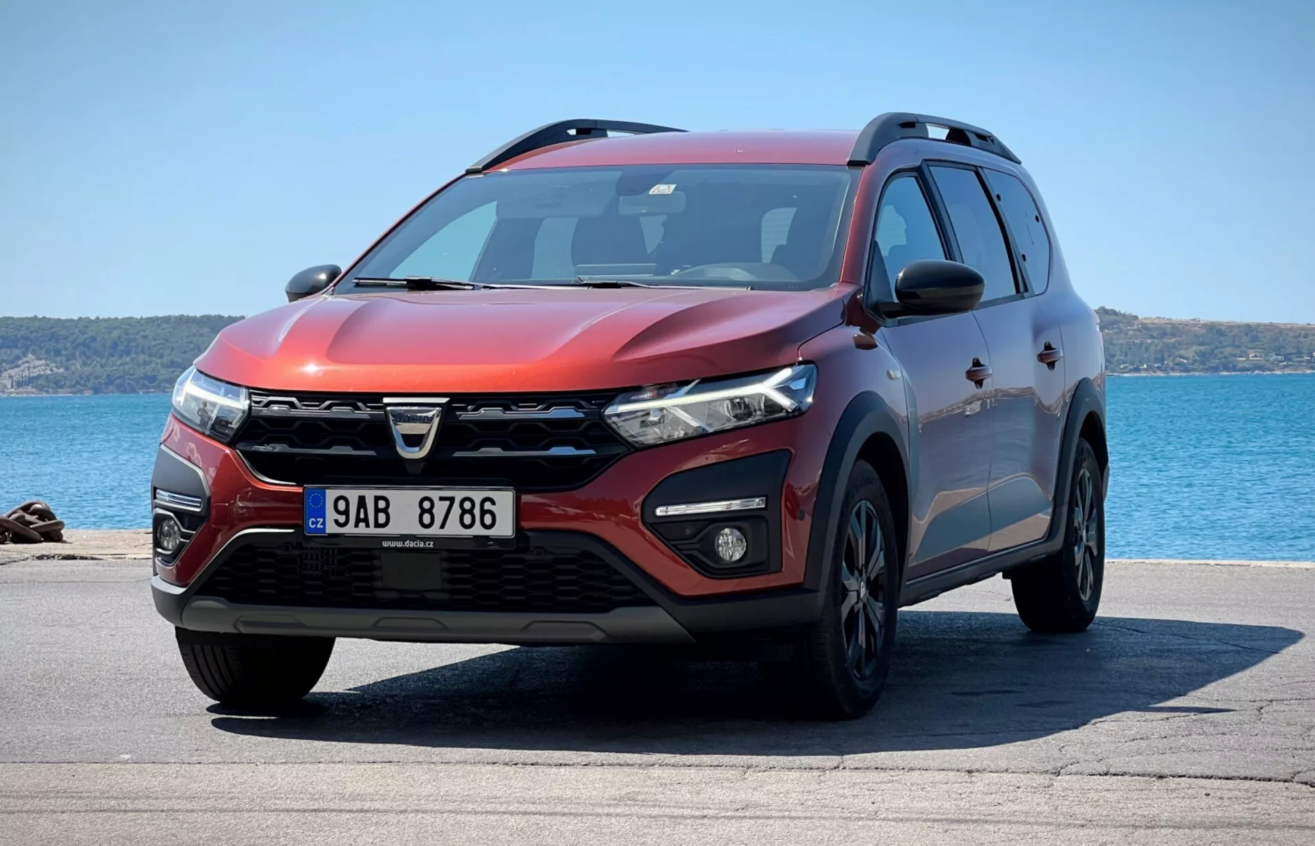 Vista frontale da tre quarti del SUV Dacia Jogger 2022 parcheggiato con sfondo oceanico, mostrando design distintivo della griglia, fari LED, luci di nebbia, paraurti anteriore aerodinamico con prese d'aria e sensori di parcheggio, contorni muscolosi del cofano, ruote in lega, segnali di svolta integrati sugli specchietti laterali, profilo laterale con protezione inferiore, barre sul tetto, finestre oscurate e una esterno rosso metallico sotto la luce solare brillante che indica profilo di design, caratteristiche tecnologiche e utilità.