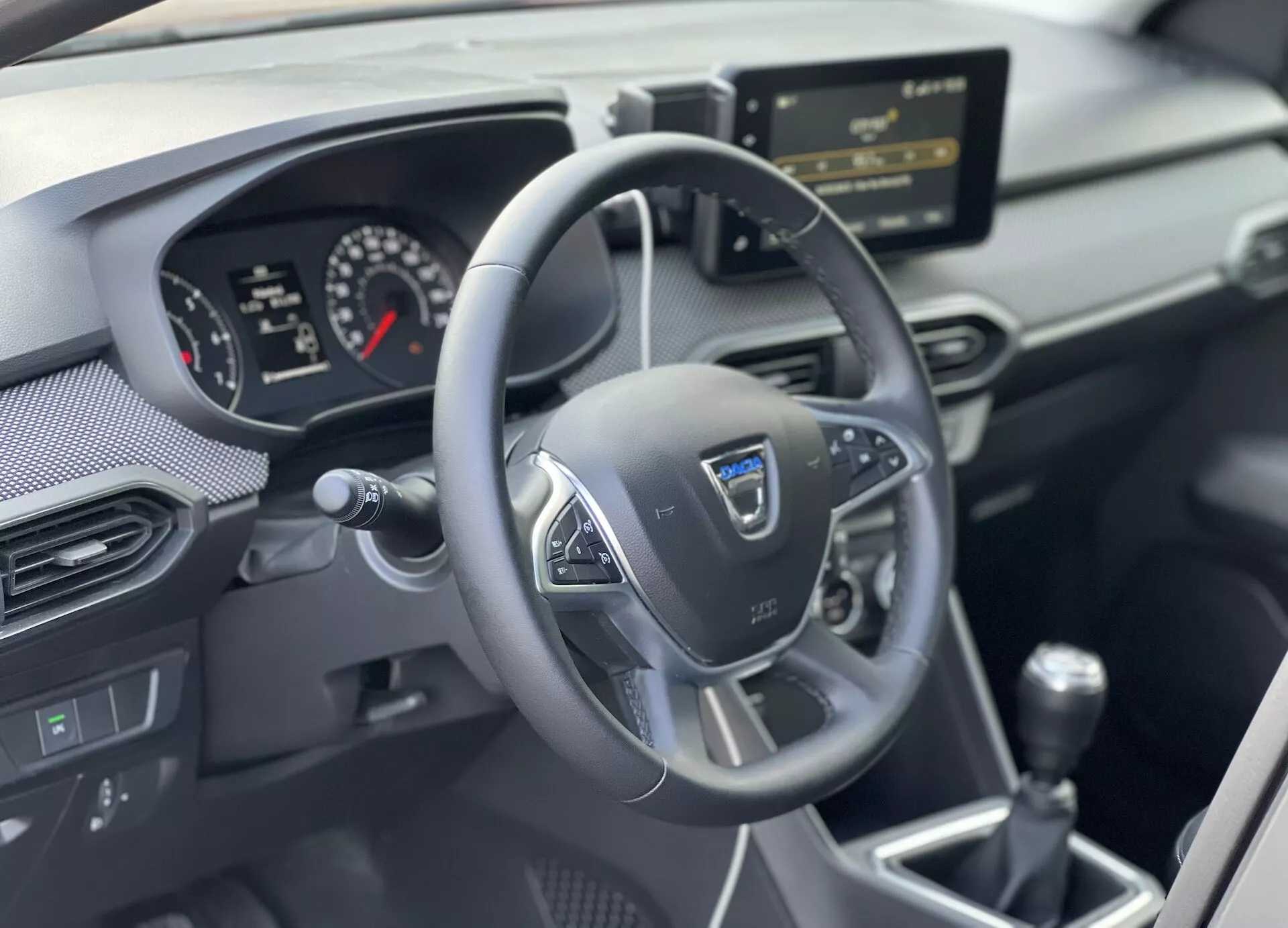 Vista interna del Dacia Jogger 2022 focalizzata sull'area del guidatore con volante multifunzione a tre razze, cruscotto analogico-digitale, console centrale con sistema di infotainment e leva del cambio manuale