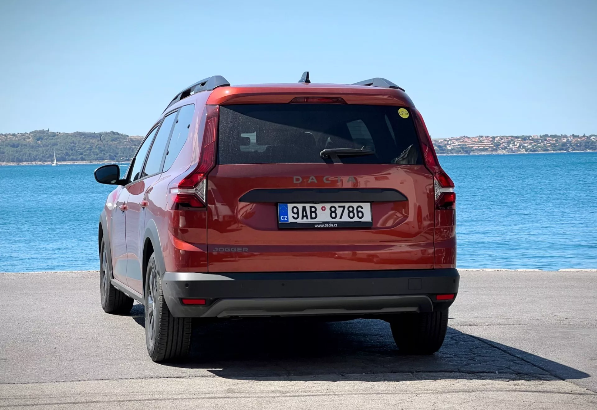 Vista posteriore di un SUV compatto rosso Dacia Jogger 2022 parcheggiato su un'area pavimentata in riva al mare sotto la luce brillante del giorno, mostrando l'intera parte posteriore del veicolo inclusi fanali posteriori, lunotto e targa contro uno sfondo di acqua blu e cieli sereni.