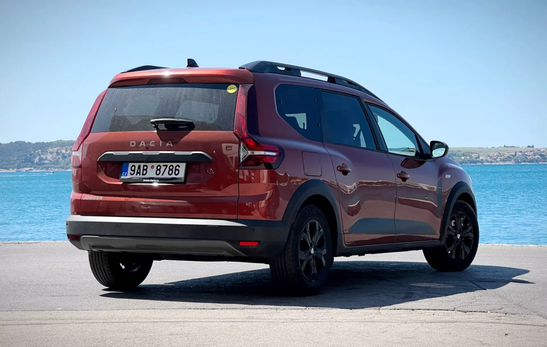 Dacia Jogger 2022 fotografata con una vista posteriore di tre quarti posizionata su una superficie pavimentata con un corpo d'acqua e cielo sereno sullo sfondo, mostrando luci posteriori moderne, un distintivo Dacia posizionato centralmente e un profilo laterale elegante contro un tranquillo ambiente costiero o lacustre.