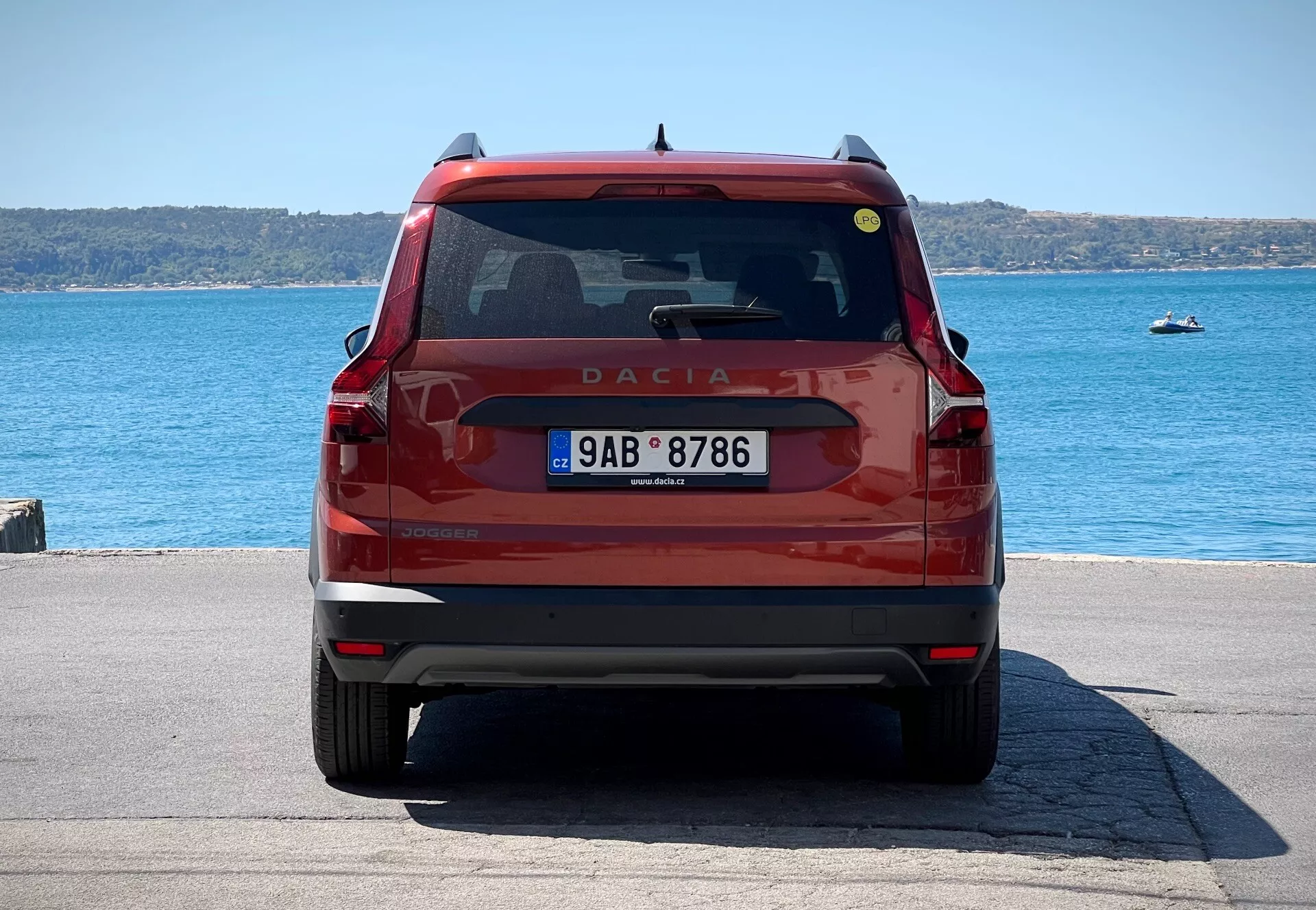 Vista posteriore di una Dacia Jogger 2022 di colore rosso, con dettagli del portellone con emblema e nome del modello, lunotto posteriore con tergicristallo e luce di stop, fanali posteriori moderni, targa e paraurti con riflettori e potenziali sensori di parcheggio, parcheggiata in riva al mare alla luce del giorno.