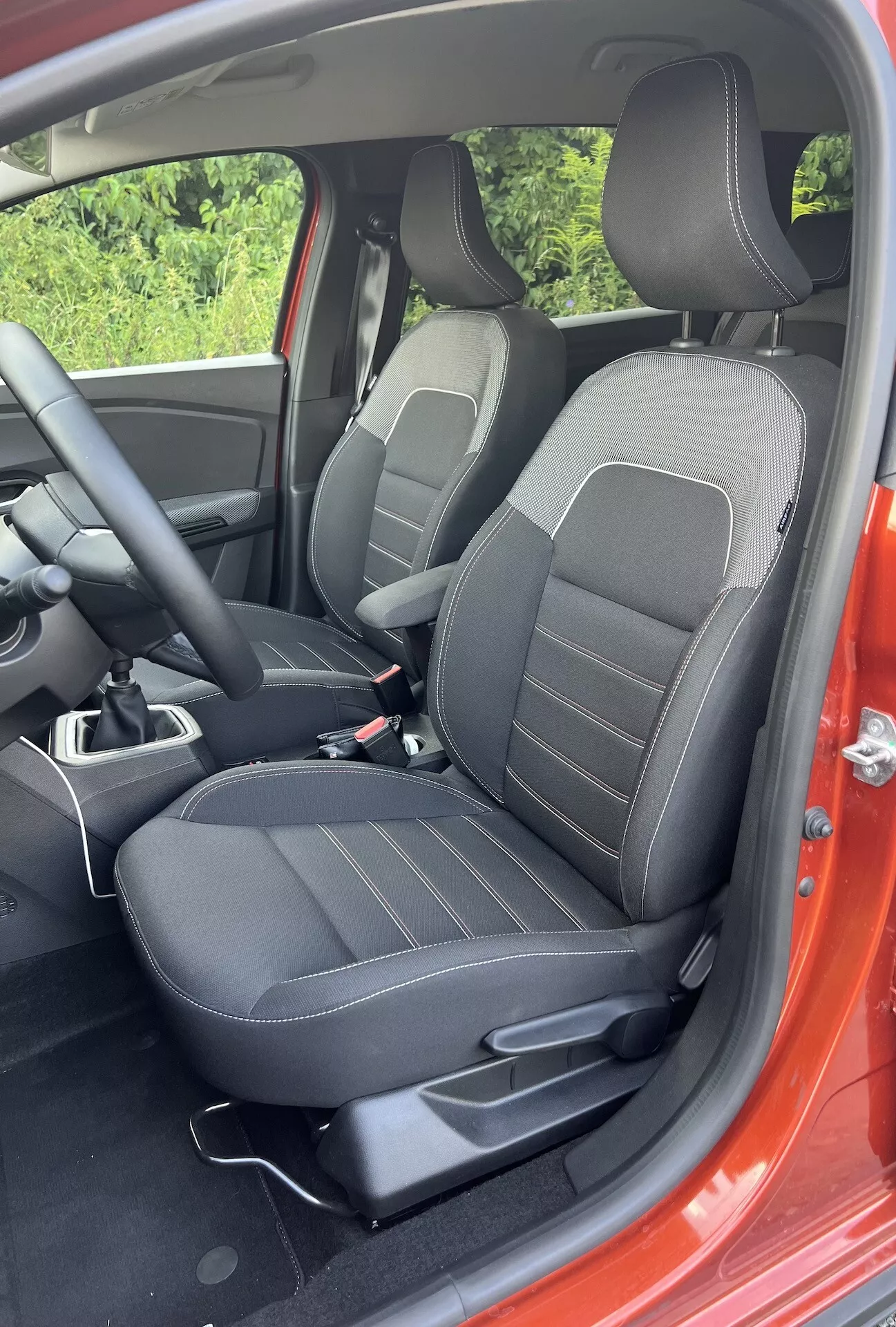 Vista dettagliata degli interni del Dacia Jogger 2022 che mostra l'area del passeggero anteriore con sedili rivestiti in nero con motivi a linee, controlli di regolazione, parte della console centrale e un accenno dell'esterno rosso