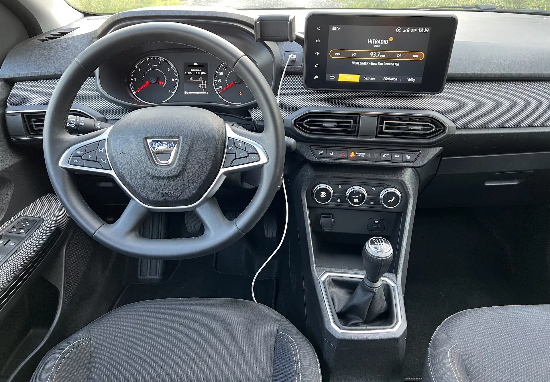 Vista interna di una Dacia Jogger 2022 dal sedile posteriore, mettendo in evidenza il cruscotto del conducente, il volante con logo del produttore e pulsanti di controllo, il quadro strumenti digitale-analogico, il sistema di infotainment touch, i controlli HVAC, il controllo climatico bizona e la leva del cambio manuale