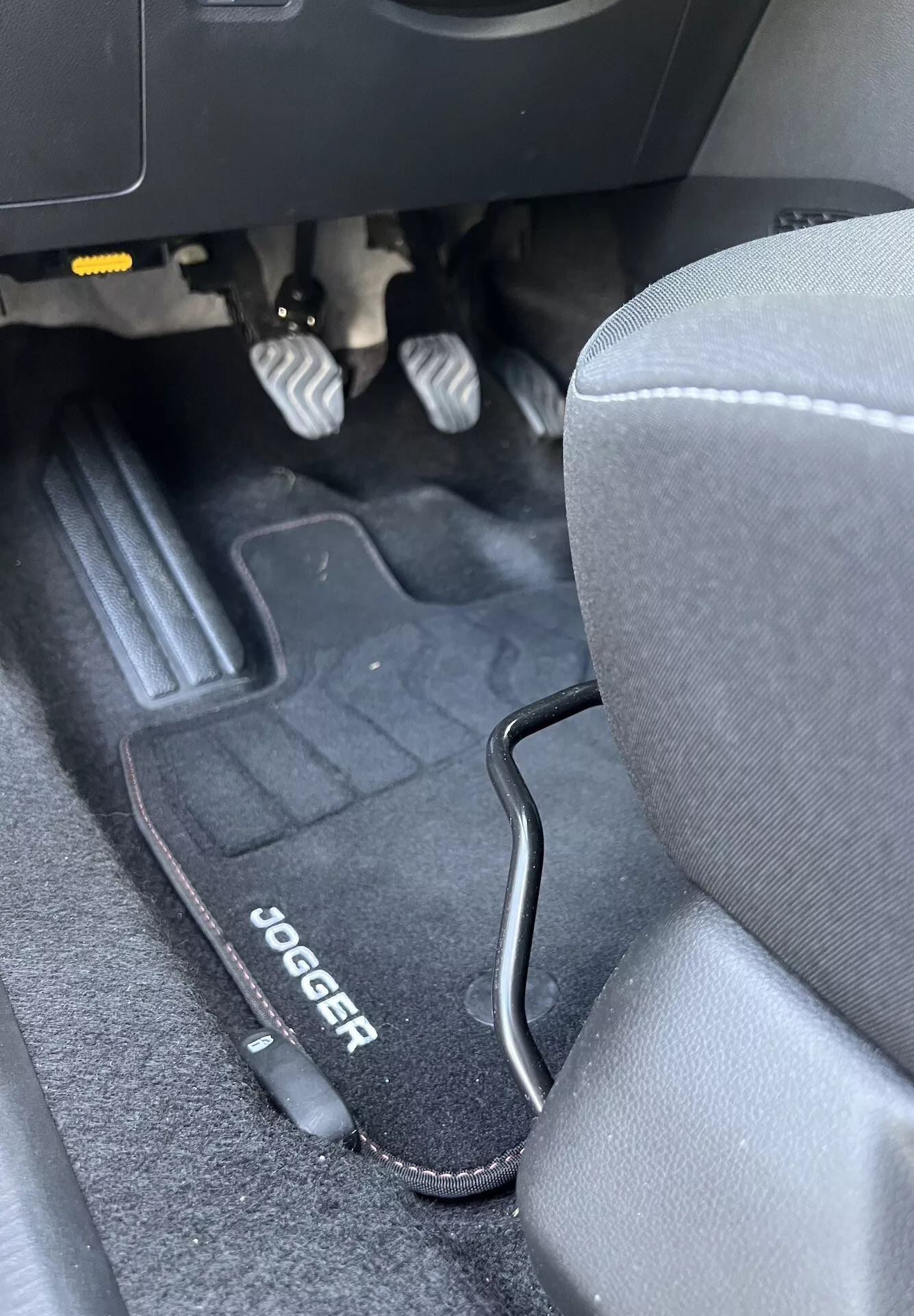 Interno della Dacia Jogger 2022 che mostra il vano piedi lato guidatore con pedali metallici, tappetino di marca, sedile in tessuto grigio, e bordo in plastica nera