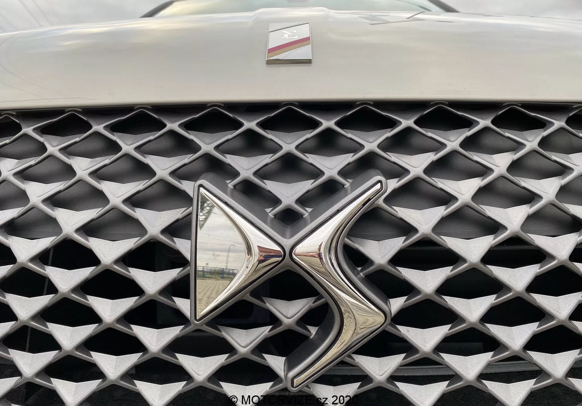 Primo piano della griglia della DS Automobiles DS 3 Crossback E-Tense 2019. L'immagine cattura la griglia da una prospettiva frontale, concentrandosi su un emblema metallico altamente riflettente. La griglia presenta una texture distintiva con celle a forma di diamante in una finitura grigia opaca, in contrasto con l'emblema cromato lucido. Campo visivo limitato oltre la griglia e l'emblema, con uno sfondo sfocato.