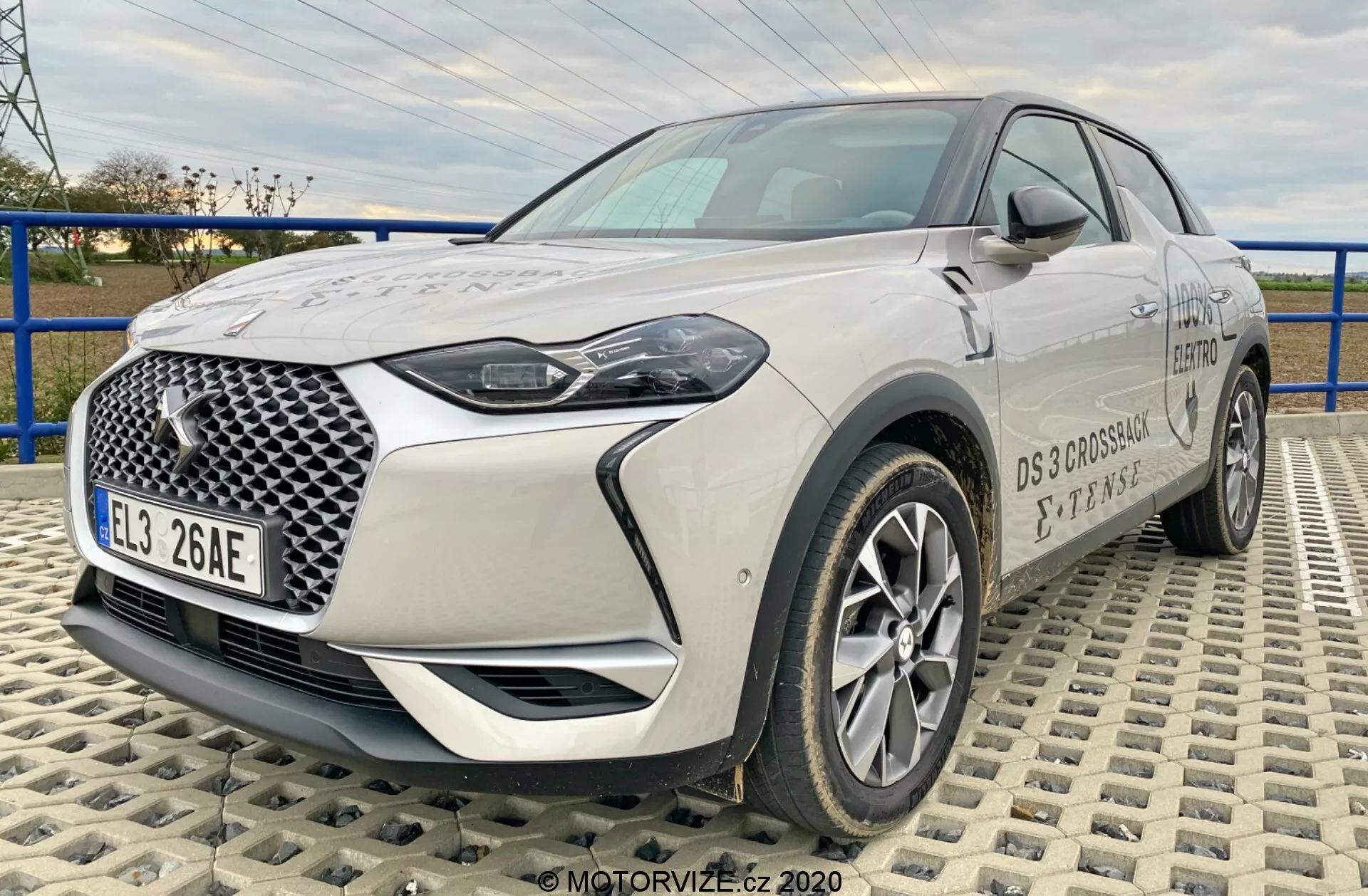 L'immagine mostra l'SUV crossover moderno DS-Automobiles DS-3 2019 (DS-3 Crossback E-Tense), catturato da un'angolazione di tre quarti frontale su una superficie strutturata realizzata con piastrelle ad incastro. La prospettiva da un'altezza medio-bassa enfatizza la statura dell'auto, fornendo una vista completa della griglia frontale con un motivo geometrico distintivo, fari a LED, cerchi in lega, specchietti laterali e dettagli del marchio. La lunghezza e le caratteristiche del design frontale del veicolo sono chiaramente visibili, evidenziando la sua tecnologia automobilistica avanzata e moderna.