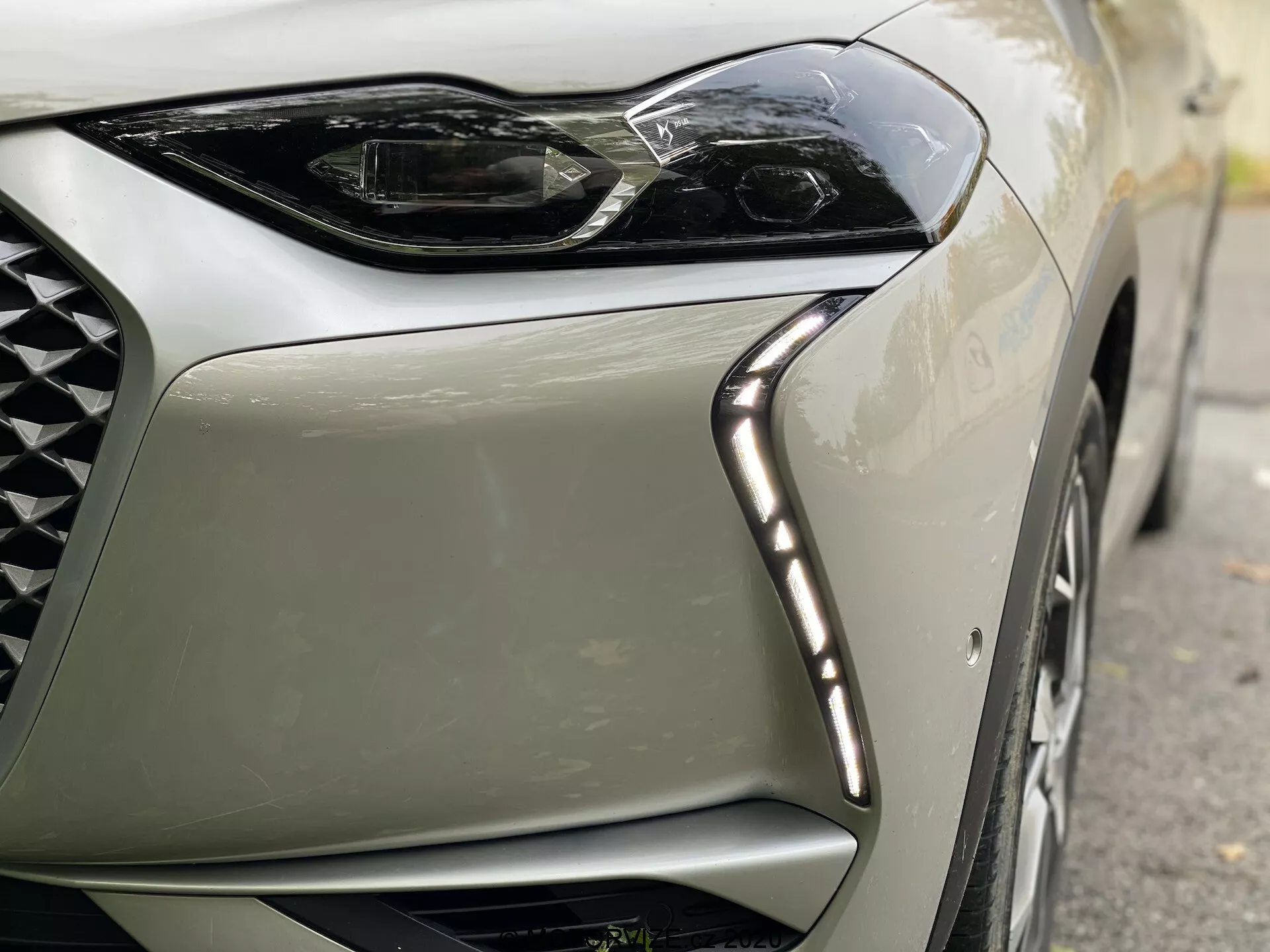 Vista ravvicinata dell'angolo anteriore sinistro di una moderna DS Automobiles DS-3 Crossback E-Tense del 2019. L'immagine si concentra sul design angolare e elegante del faro a LED, con luci diurne a LED lungo il bordo superiore e parte della griglia a rete con disegno a diamante, enfatizzando lusso e tecnologia all'avanguardia.