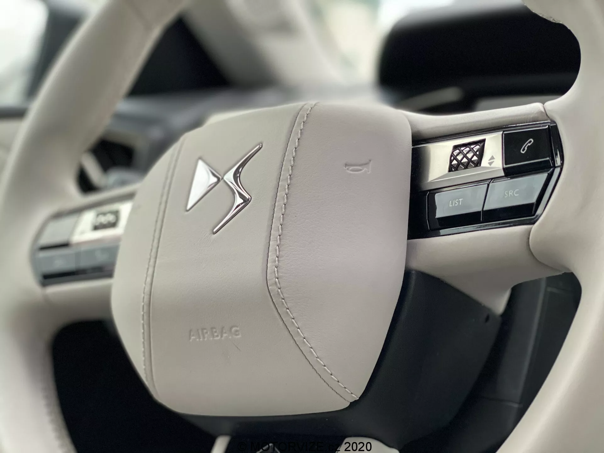 Primo piano del lato sinistro di un volante moderno rivestito in pelle chiara della DS Automobiles DS 3 Crossback E-Tense 2019, che enfatizza la maestria e il lusso con cuciture visibili. Dispone di diversi pulsanti integrati per il controllo delle funzioni del veicolo, compresi comandi vocali, telefono, menu principale e controlli multimediali, accanto a un emblematico centrale prominente e una custodia 'AIRBAG' in rilievo.