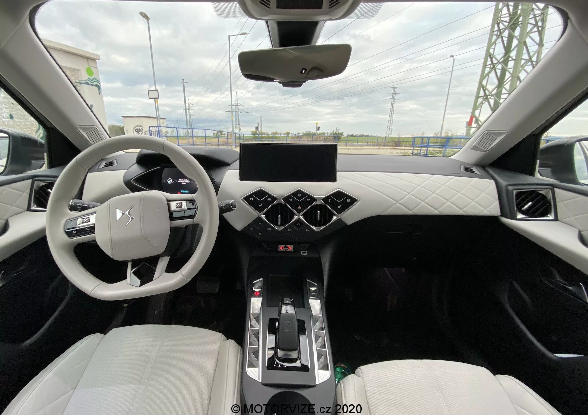Vista dal sedile posteriore di una DS Automobiles DS-3 Crossback E-Tense del 2019 che mostra il moderno interiore centrale. La foto cattura un volante a tre razze con comandi montati, un pannello strumenti digitale, un grande schermo touchscreen sulla console centrale con pulsanti esagonali per il controllo del clima al di sotto e una leva del cambio che indica una trasmissione automatica. Il cruscotto e le porte presentano un design trapuntato in ecopelle o pelle, e i sedili anteriori a secchiello sono rivestiti in pelle o materiale sintetico di colore chiaro. Caratteristiche degne di nota includono l'illuminazione ambientale lungo i bordi, un tetto panoramico e bocchette d'aria visibili sul cruscotto.