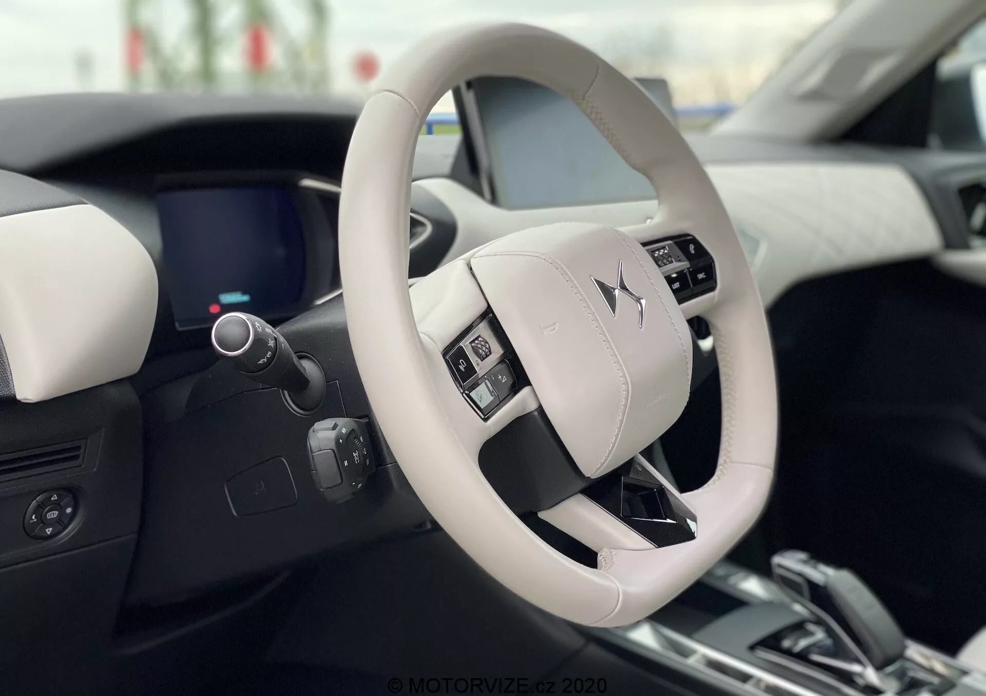 Vista interna della DS Automobiles DS 3 Crossback E-Tense 2019, che mostra il design moderno e lussuoso del volante e del cruscotto dalla prospettiva del sedile del conducente. Il volante di colore chiaro presenta pulsanti di controllo e mostra in modo prominente il logo del produttore. Il cruscotto digitale elegante, parzialmente visibile dietro il volante, completa il design interno bicolore con materiali di alta qualità.