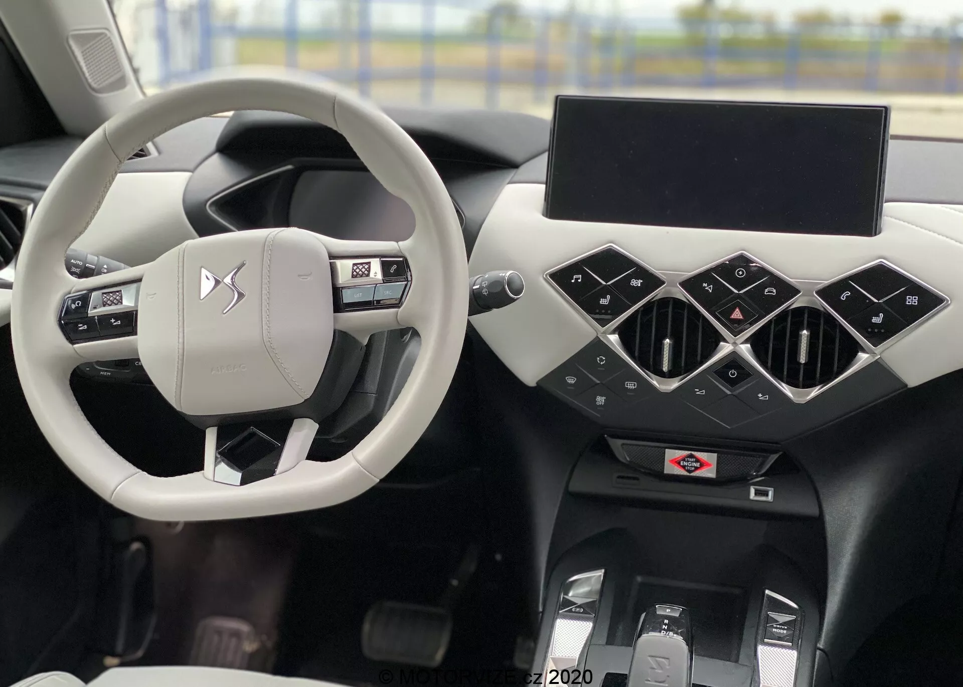 Scatto dell'interno di una DS Automobili DS 3 Crossback E-Tense del 2019 con il volante argentato/grigio chiaro al centro, con pulsanti di controllo e il logo del produttore, attraverso il quale si intravede il sedile del conducente. Il cruscotto nero presenta elementi geometrici con prese d'aria centrali triangolari, un display di infotainment spento e sotto comandi dell'aria condizionata e tunnel centrale. A sinistra si intravede la porta del conducente con comandi per i finestrini e gli specchietti.