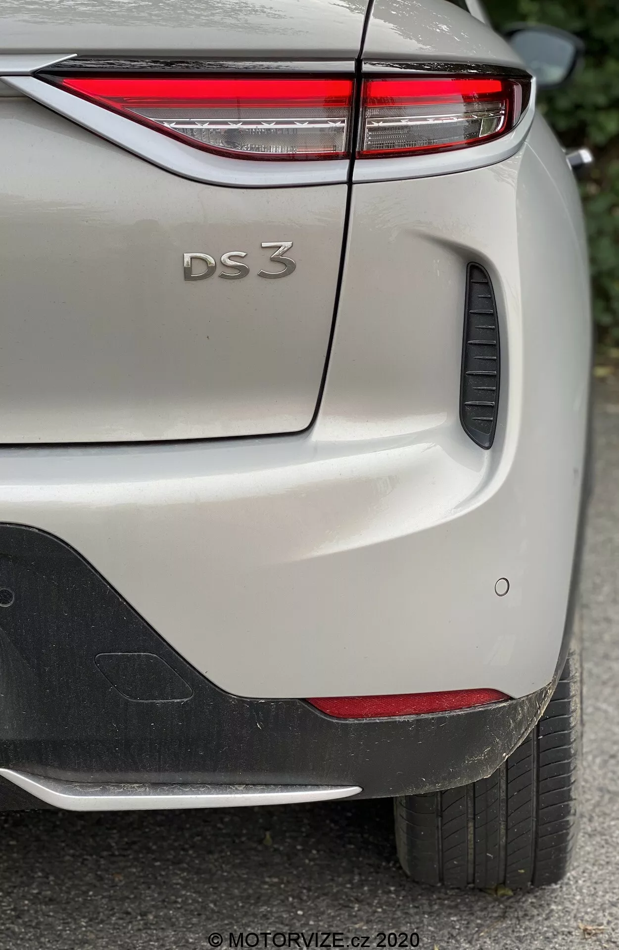 Vista laterale posteriore ravvicinata di una DS Automobiles DS 3 Crossback E-Tense 2019, con particolare attenzione alla parte superiore destra del paraurti posteriore, il portellone del bagagliaio e una parte del parafango posteriore destro. L'immagine cattura i dettagli distintivi del design del modello, comprese le sottili luci posteriori a LED, la scritta in rilievo 'DS 3', i sensori di parcheggio e una caratteristica in plastica nera sul paraurti. L'auto, mostrata in una tonalità chiara con finitura metallica, è fotografata alla luce naturale, mettendo in evidenza i dettagli fini della vernice e della struttura.