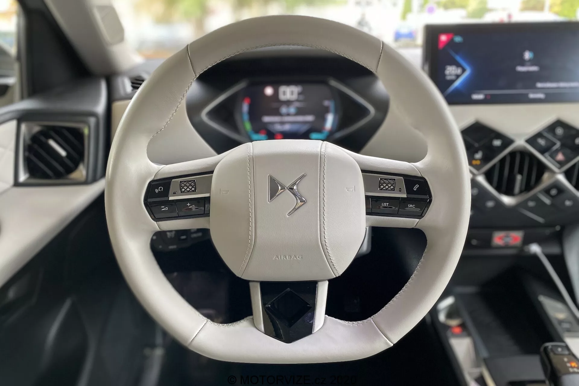 Vista interna dalla prospettiva del conducente in un DS Automobiles DS 3 Crossback E-Tense 2019, con particolare attenzione al moderno volante a due razze, rivestito in pelle chiara, dotato di controlli multifunzionali e un cruscotto digitale che visualizza velocità e giri al minuto (RPM). La console centrale vanta uno schermo touch di infotainment integrato e controlli minimalisti per il sistema di climatizzazione, evidenziando la tecnologia automobilistica avanzata e un design lussuoso.
