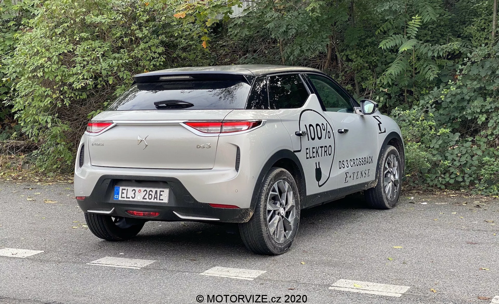 DS Automobiles DS 3 2019 (DS 3 Crossback E-Tense) catturato da un'angolazione posteriore di tre quarti, evidenziando il profilo posteriore e laterale del veicolo. Le caratteristiche del design includono fanali posteriori orientati orizzontalmente, una rifinitura in cromo, maniglie delle porte a filo, una porta di ricarica e cerchi in lega multi-razza, emblematici di un'auto elettrica moderna. L'auto è parcheggiata all'aperto in un ambiente urbano o suburbano sotto un cielo coperto, con alberi decidui nelle vicinanze.