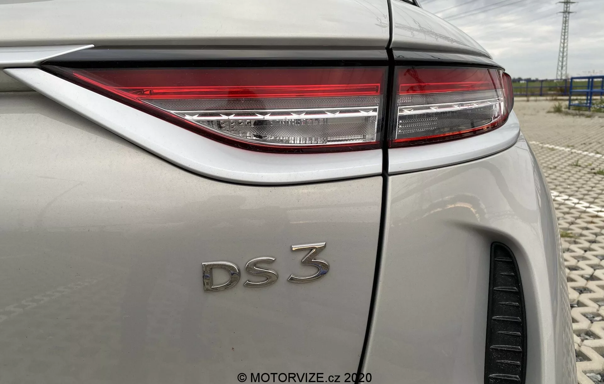 Vista ravvicinata dell'angolo posteriore a tre quarti della DS Automobiles DS 3 Crossback E-Tense 2019. L'immagine si concentra sul lato destro del veicolo, evidenziando luci posteriori a LED nitide, il badge 'DS 3' e una carrozzeria metallica leggera. Le caratteristiche visibili includono la linea di piega del parafango posteriore, la parte del pneumatico posteriore, l'antenna a pinna di squalo e la transizione del lunotto nella linea del tetto, tutto sotto il riflesso di un cielo nuvoloso.