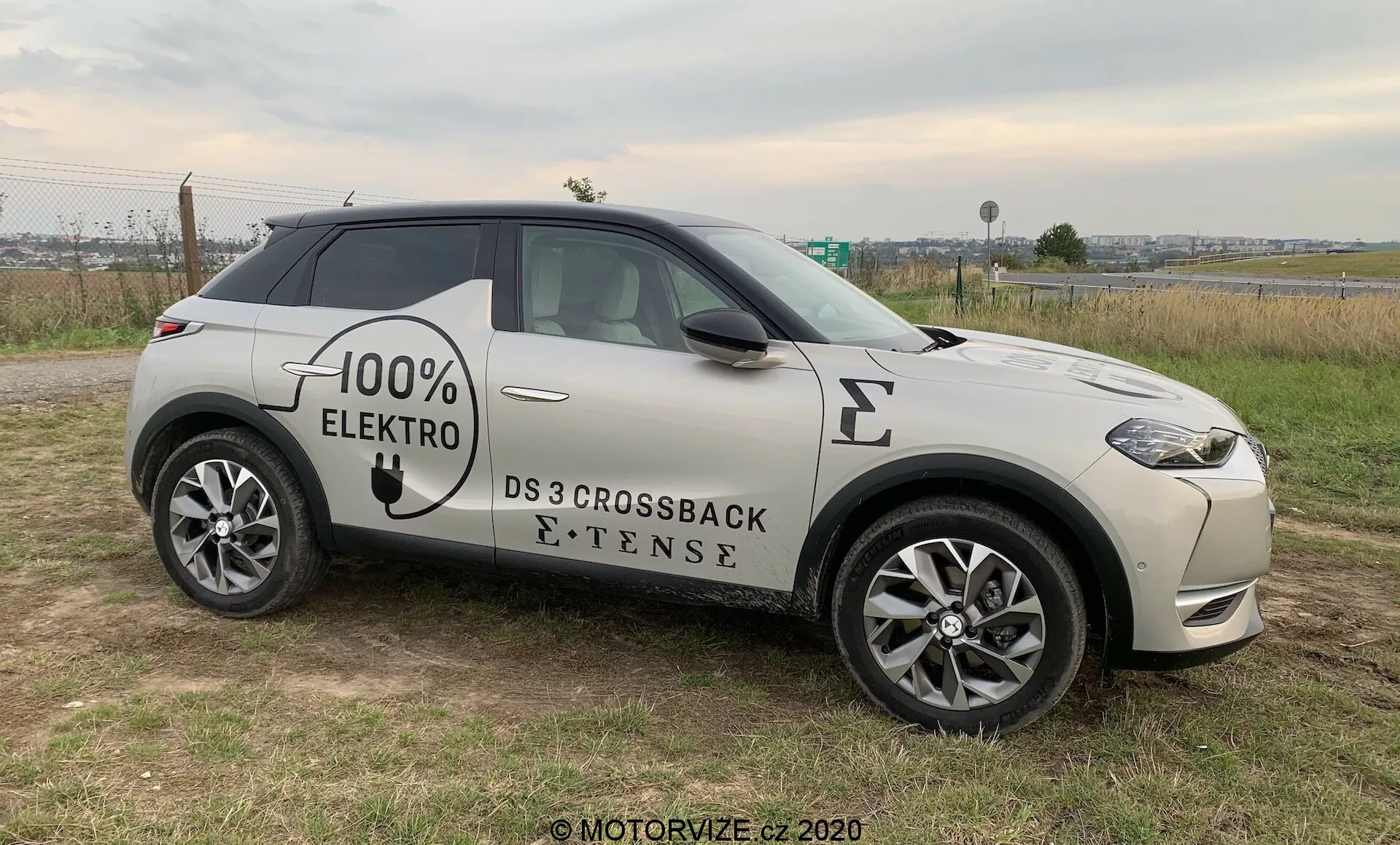 Vista di profilo laterale di un SUV moderno, il DS 3 Crossback E-Tense del 2019, un veicolo elettrico con grafica '100% ELEKTRO' sul lato. Posizionato su terreno erboso con un po' di strada asfaltata sullo sfondo, che permette una chiara visione della griglia anteriore, fari LED, ruota anteriore e profilo da un angolo leggero. Le caratteristiche visibili includono cerchi in lega a più razze, specchietti laterali con indicatori integrati, maniglie delle porte a filo e simbolo della presa di ricarica elettrica sul parafango anteriore, suggerendo un'attenzione alla tecnologia avanzata, allo stile e all'eco-sostenibilità, il tutto sotto una luce naturale e nuvolosa.