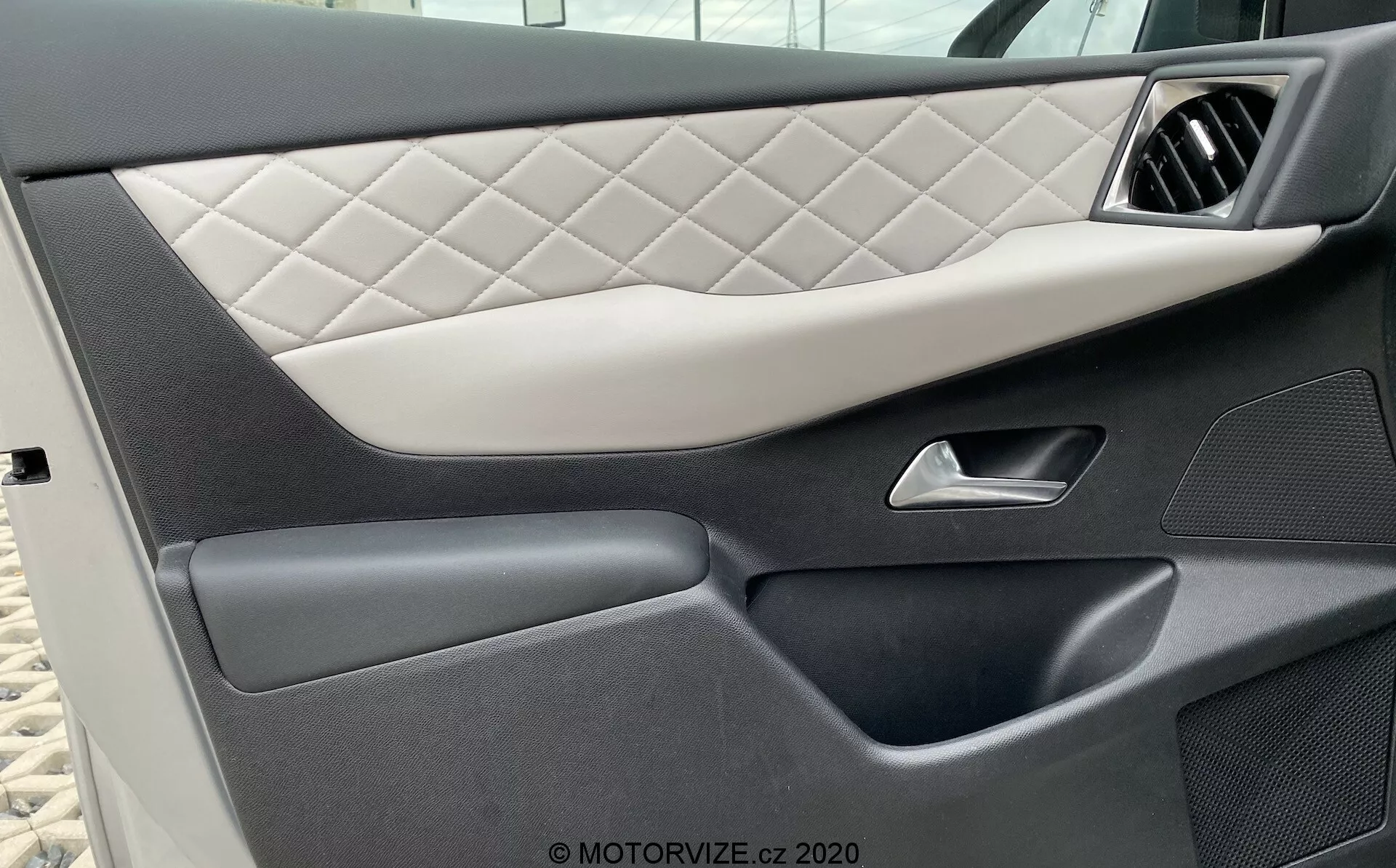Vista ravvicinata del pannello della porta interna del veicolo DS Automobiles DS-3 2019 (DS-3-Crossback-E-Tense), che mostra l'imbottitura trapuntata con motivo a diamante, una maniglia argentata elegante, sezioni in plastica scura di alta qualità con trame diversificate e una tasca portaoggetti inferiore senza soluzione di continuità, evidenziando il design lussuoso e dettagliato dell'auto.