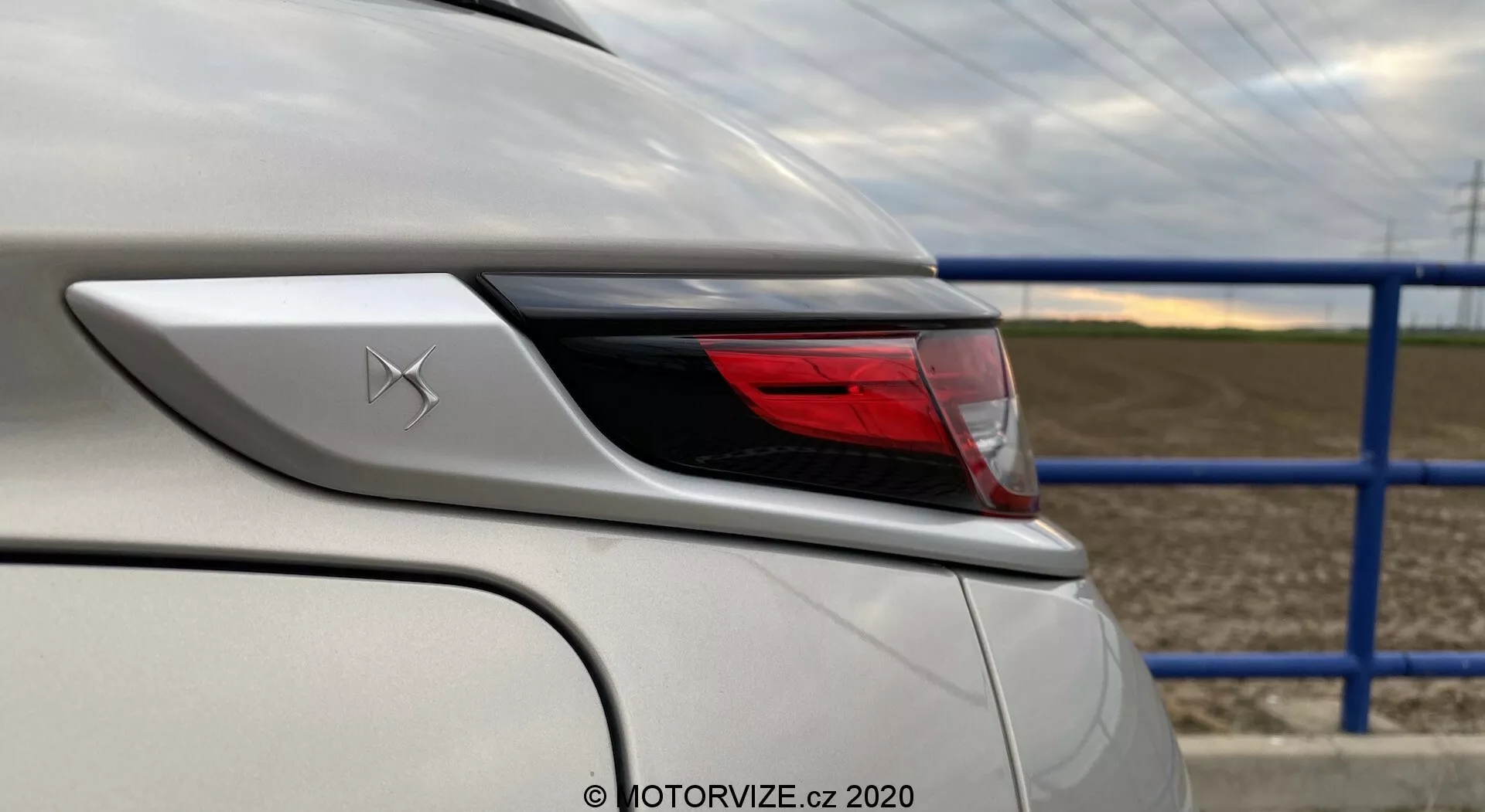 Primo piano dell'angolo posteriore di un veicolo bianco DS-3 2019 (DS-3 Crossback E-Tense), focalizzato sugli elementi di design e le caratteristiche attorno all'area del fanale posteriore. L'inquadratura dall'angolo posteriore sinistro evidenzia la curvatura del fanale posteriore e del parafango posteriore, mostrando dettagli della carrozzeria dell'auto e l'avanzato assemblaggio delle luci a LED. Un distintivo con una X stilizzata è visibile sopra il fanale posteriore, con la vernice bianca in perfette condizioni che riflette una finitura lucida. Sullo sfondo c'è una strada con una barriera metallica blu e un campo agricolo.