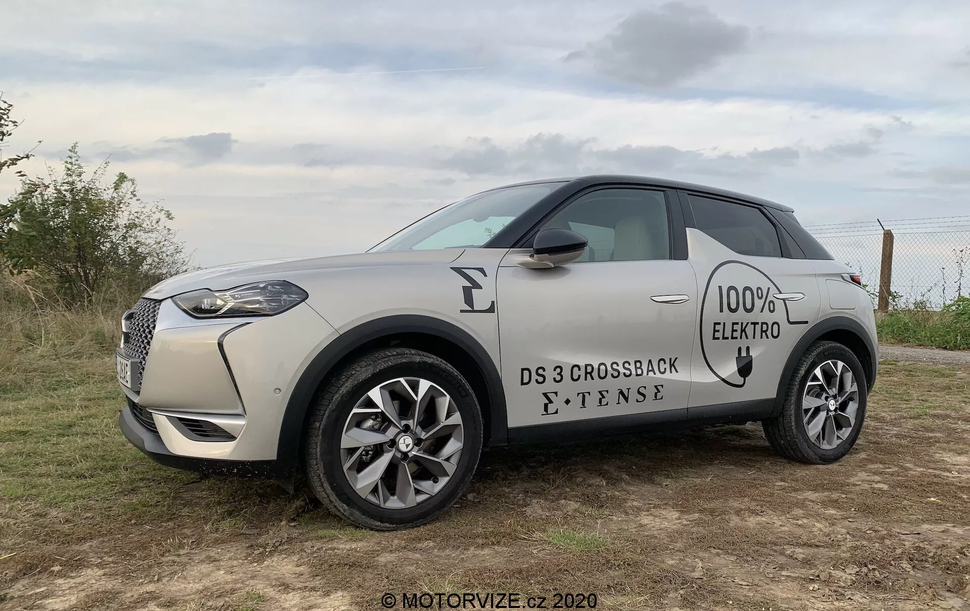 Profilo laterale di un SUV compatto elettrico DS 3 Crossback E-Tense 2019 in una tonalità sabbia chiara con tetto nero a contrasto. L'immagine cattura la griglia anteriore con cornice cromata, fari a LED, distintivi '100% ELECTRO' e 'E-TENSE' sul lato, cerchi in lega a più raggi e maniglie a filo. Il veicolo è posizionato all'aperto su un terreno erboso sotto un cielo nuvoloso, suggerendo illuminazione di prima mattina o tardo pomeriggio.