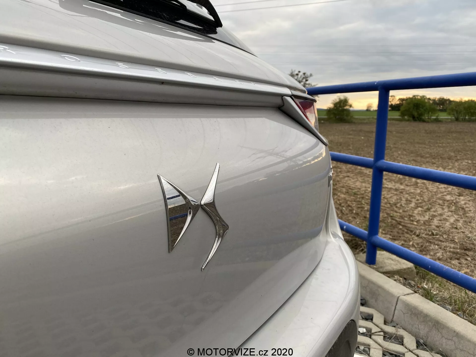 Primo piano della sezione posteriore di una berlina o hatchback di lusso di colore argento chiaro o grigio, con un prominente emblema del marchio sul bagagliaio e un moderno gruppo ottico posteriore a LED. La fotografia, scattata con un leggero angolo da vicino alla parte posteriore verso il lato destro del veicolo, mostra linee di design orizzontali nette e uno sfondo suburbano o rurale con ringhiere blu e strutture. L'estetica del design dell'auto mette in risalto la tecnologia contemporanea e uno schema di colori neutri e sobri.