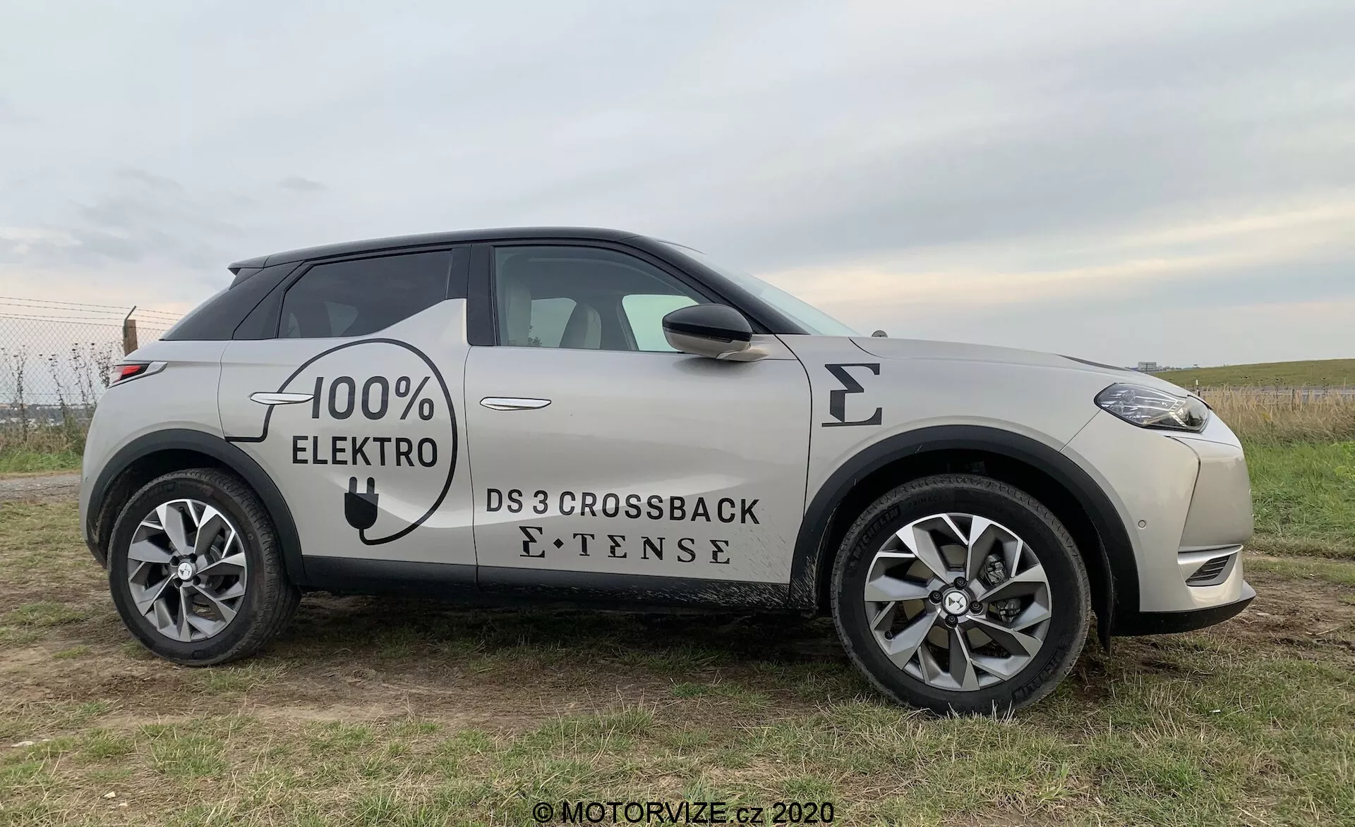 Profilo laterale della DS Automobiles DS 3 Crossback E-Tense 2019, un veicolo elettrico, fotografato da un leggero angolo frontale, che mette in risalto la ruota anteriore, la portiera lato guida e il retrovisore, con viste parziali della griglia frontale e della ruota posteriore, posizionato su un campo erboso con uno sfondo sfumato.