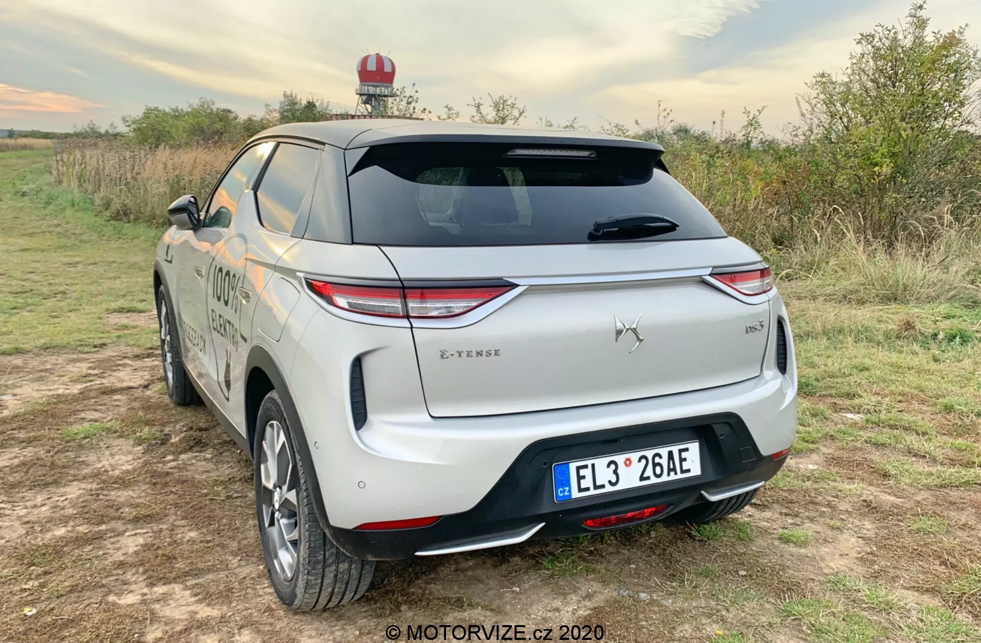 Vista posteriore tre quarti di un SUV DS Automobiles DS-3 Crossback E-Tense del 2019 parcheggiato su una superficie non asfaltata in un contesto rurale, che mette in mostra luci posteriori a LED, emblema ben visibile, tergicristallo posteriore, paraurti posteriore distintivo con sensori, finitura bicolore, targa centrale, distintivo del livello di allestimento, portapacchi, possibile luce di emergenza, linea del corpo aerodinamica liscia, ruote in lega, specchietti laterali dello stesso colore della carrozzeria e uno sportellino di ricarica che indica che si tratta di un veicolo elettrico o ibrido plug-in.