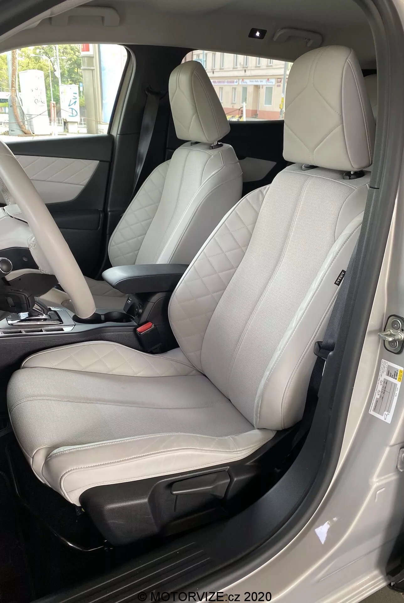 Interni della DS Automobiles DS 3 Crossback E-Tense 2019, focalizzati sul sedile del conducente da una prospettiva esterna del lato del conducente. L'immagine, scattata da un angolo basso verso alto, mostra un moderno sedile in pelle bicolore con centro grigio chiaro e bordi grigio scuro o neri. Chiaramente progettato per comfort e estetica sportiva, il sedile ha imbottiture laterali prominenti, cuciture visibili e controlli di regolazione elettronica. La porta del conducente aperta, la cintura di sicurezza visibile, la console centrale parzialmente visibile e il pannello della porta con vari pulsanti di controllo indicano materiali premium e molteplici funzioni avanzate. L'illuminazione diurna migliora la chiarezza dei dettagli dell'interno di lusso.
