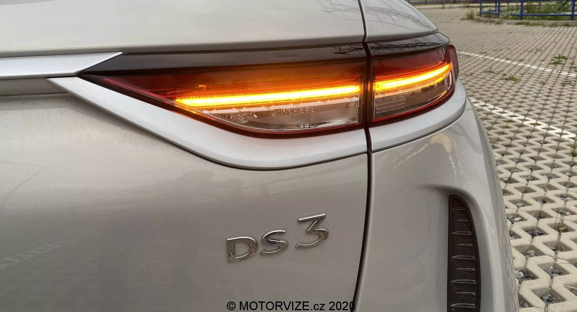 Primo piano della sezione posteriore di una DS 3 Crossback E-Tense (modello 2019), che mostra il quarto posteriore destro da un angolo leggermente elevato. L'immagine mette in evidenza le luci posteriori a LED e la freccia con illuminazione arancione brillante. Gli elementi visibili includono il bordo superiore del bagagliaio, la curvatura del parafango posteriore, l'allineamento preciso dei pannelli del corpo e parte della ruota posteriore destra. L'auto è di colore argento o grigio chiaro. La foto è stata scattata su una superficie testurizzata.