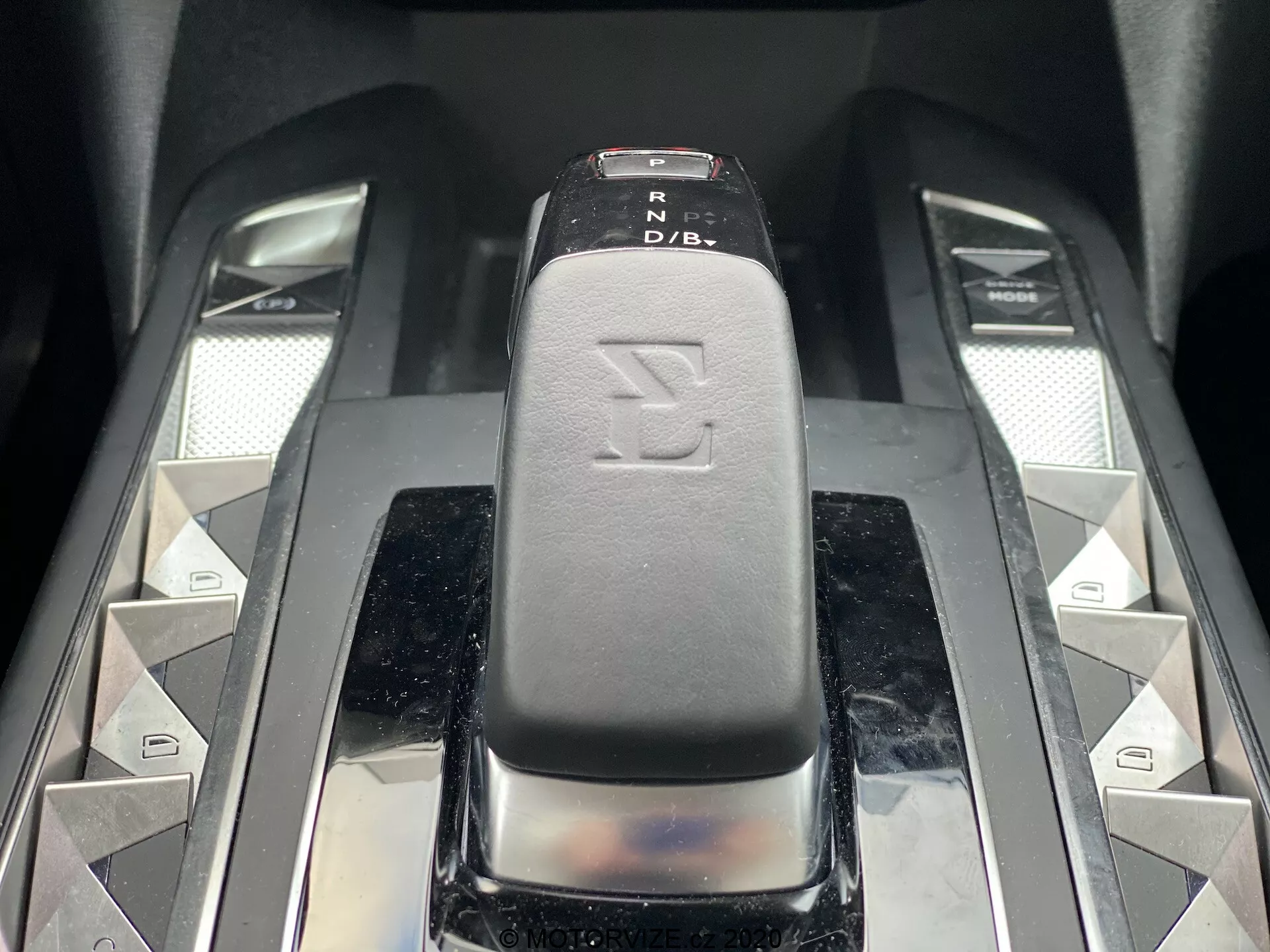 Primo piano della leva del cambio in una DS Automobiles DS-3 2019 (DS-3 Crossback E-Tense), catturato da un angolo leggermente elevato anteriore. La leva del cambio, con una superficie in pelle o simile pelle nera con indicatori PRND, è il punto centrale dell'immagine. Pulsanti simmetrici a forma di diamante argentato e due cursori metallici affiancano la leva, suggerendo funzionalità aggiuntive dell'auto. I materiali ad alta lucentezza e di alta gamma trasmettono un interno moderno e lussuoso.