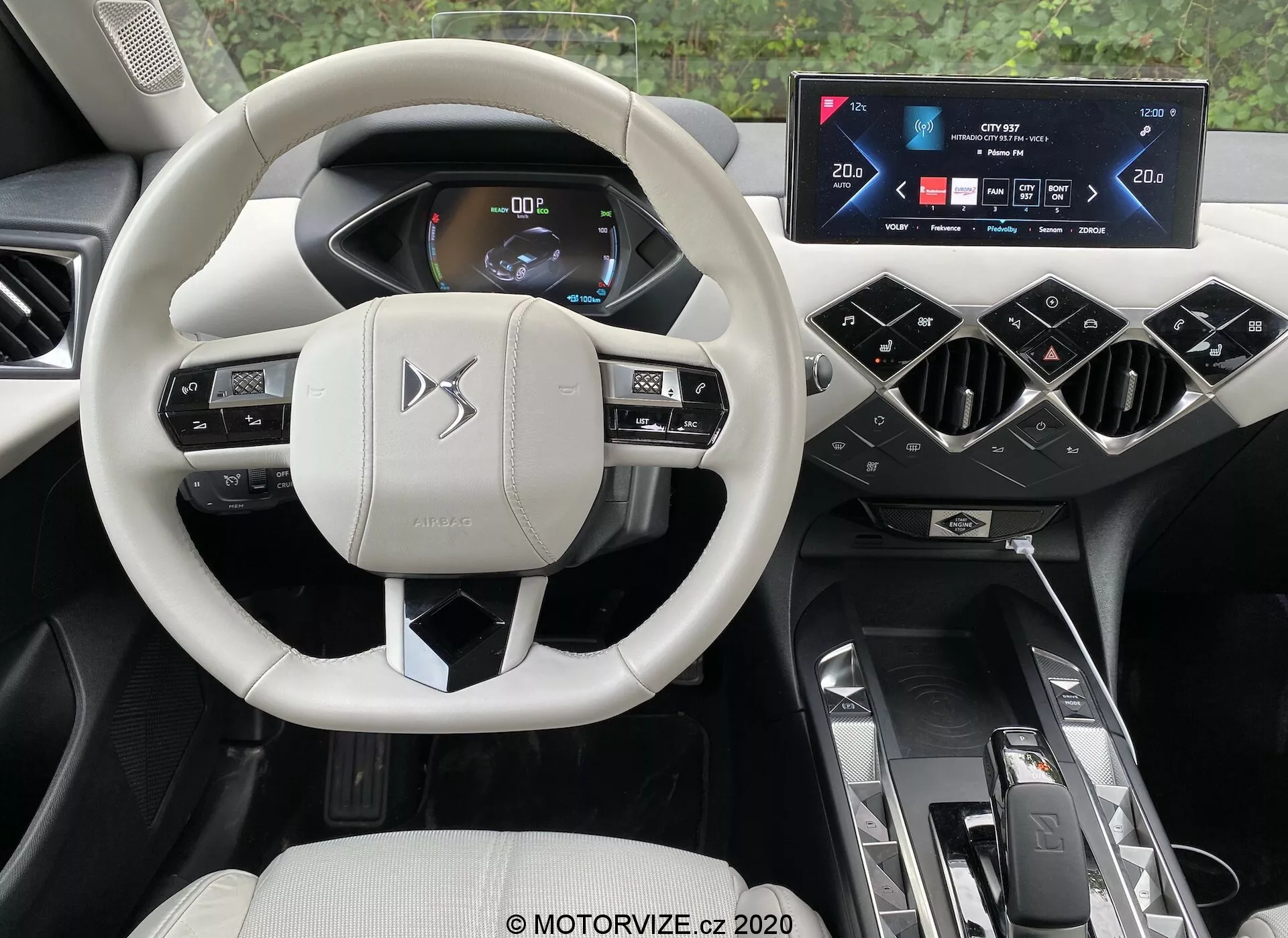 Vista dell'interno di un'auto moderna dalla prospettiva del conducente, evidenziando gli elementi sofisticati del ds-automobili ds-3 2019 (ds-3-crossback-e-tense). Al centro della scena un volante multifunzionale di colore chiaro con pulsanti di controllo e leve, su uno sfondo di un quadro strumenti digitale che mostra informazioni sul veicolo con grafici chiari e luminosi. Sopra la console centrale troviamo un display widescreen touchscreen dell'infotainment, incorniciato da bocchette di ventilazione angolate. Il tunnel centrale ospita un selettore del cambio automatico, controlli dei modali di guida e portabicchieri doppi. L'interno di lusso e di qualità premium presenta una combinazione di tonalità chiare e scure, che incarnano comfort, comodità e tecnologia automobilistica avanzata.