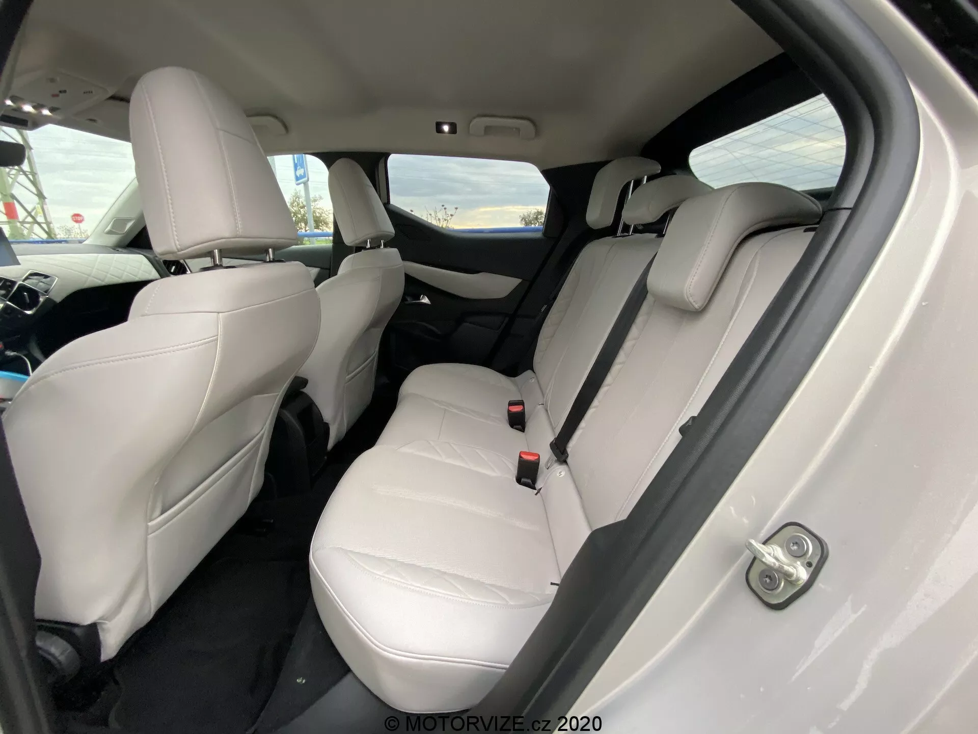 Vista interna della DS Automobiles DS 3 Crossback E-Tense 2019 da un'angolazione obliqua, concentrata sul compartimento posteriore dei passeggeri. I sedili in pelle chiara, i poggiatesta visibili, le cinture di sicurezza e un bracciolo centrale ribaltabile sul sedile posteriore indicano un interno lussuoso e ben curato. I tappetini scuri contrastano con i rivestimenti chiari e i controlli dei finestrini elettrici sono evidenti sulla porta. L'angolo della fotocamera leggermente verso il basso dalla porta posteriore destra mette in evidenza l'area dei sedili e lo spazio per le gambe.