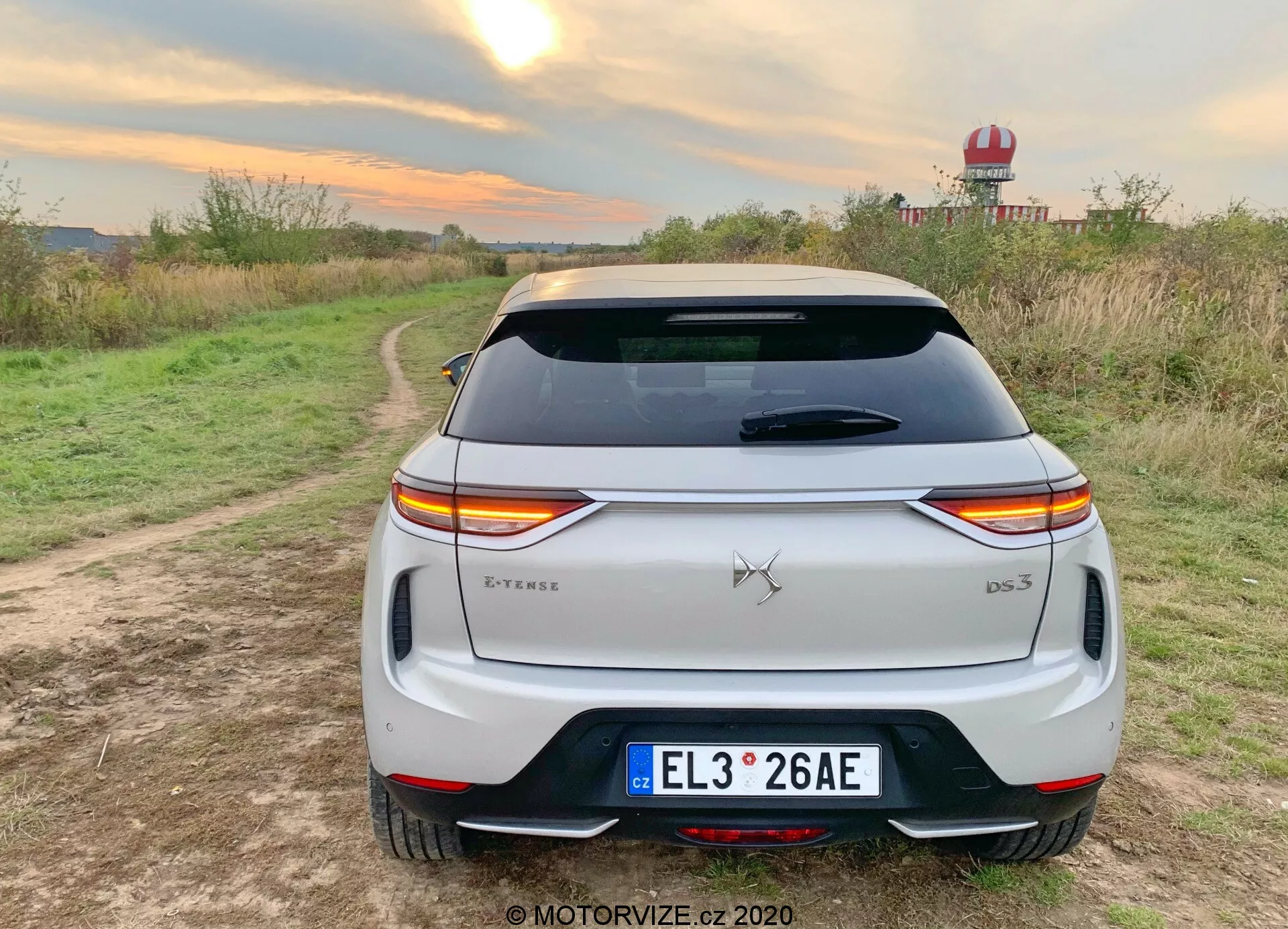 Vista posteriore di un SUV crossover bianco 2019 DS Automobiles DS 3 Crossback E-Tense parcheggiato su un sentiero sterrato con vegetazione su entrambi i lati in un ambiente rurale. I distintivi fanali posteriori a LED si estendono lungo la parte posteriore, interrotti dal emblema del marchio. Il badge del modello 'DS 3' si trova in basso a destra del bagagliaio, con un badge 'E-Tense' in basso a sinistra che indica un motore elettrico o ibrido. Le caratteristiche includono una targa in stile europeo, un lunotto posteriore con linee di sbrinamento, uno spoiler sul tetto, un paraurti posteriore con riflettori integrati e una modanatura nera, e doppi terminali di scarico, che suggeriscono un design più sportivo. L'ambiente suggerisce un giro di prova o una sessione fotografica in stile di vita che enfatizza l'uso all'aperto, con condizioni di luce calda che accentuano la vernice bianca del veicolo.