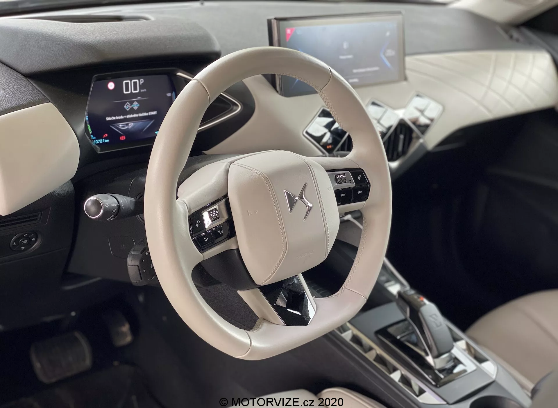 Scatto dell'interno del DS-Automobiles DS-3 2019 (DS-3 Crossback E-Tense) che si concentra sul volante beige/chiaro con emblema Mitsubishi e la console centrale. Foto scattata dalla prospettiva dei sedili posteriori, mostrando i comandi per audio, voce e sistemi di controllo della velocità. Quadro strumenti digitale che mostra una velocità pari a zero e leva del cambio in posizione 'P', indicando che l'auto è ferma. Sistema di infotainment centrale con display touchscreen visibile in un interno bicolore con accenti metallici, suggerendo display digitali moderni e caratteristiche automobilistiche avanzate.