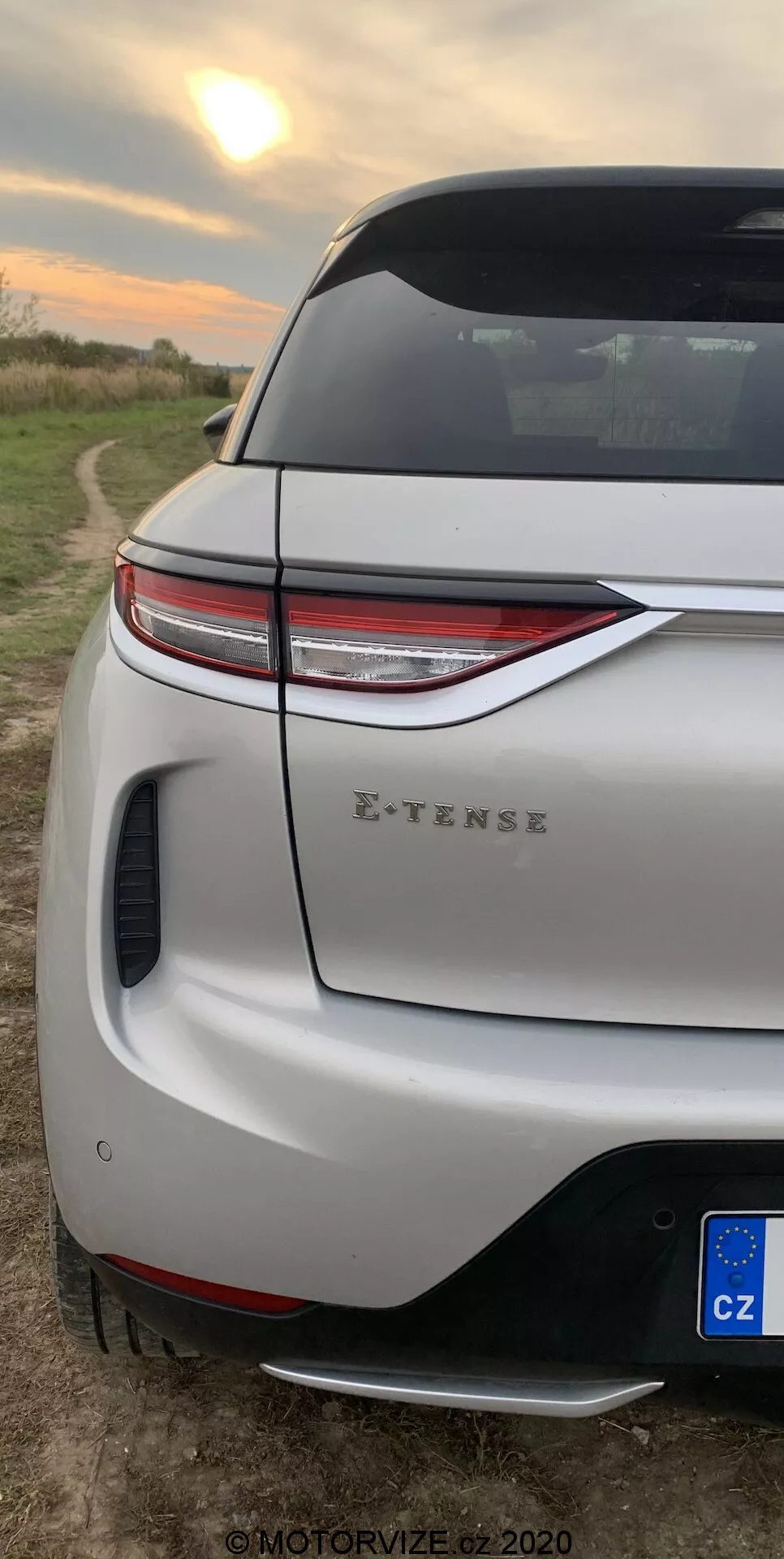 Primo piano verticale della sezione angolare posteriore della DS-3 2019 (DS-3 Crossback E-TENSE). Si concentra sui moderni fanali posteriori a LED, il paraurti posteriore sportivo con diffusore integrato e sensore di parcheggio, il portellone con spoiler aerodinamico e una visuale parziale della ruota posteriore e del parafango. Ambientato in un contesto rurale con un campo aperto e un cielo che suggerisce l'alba o il tramonto, indicando il design moderno del veicolo e le sue capacità elettriche.