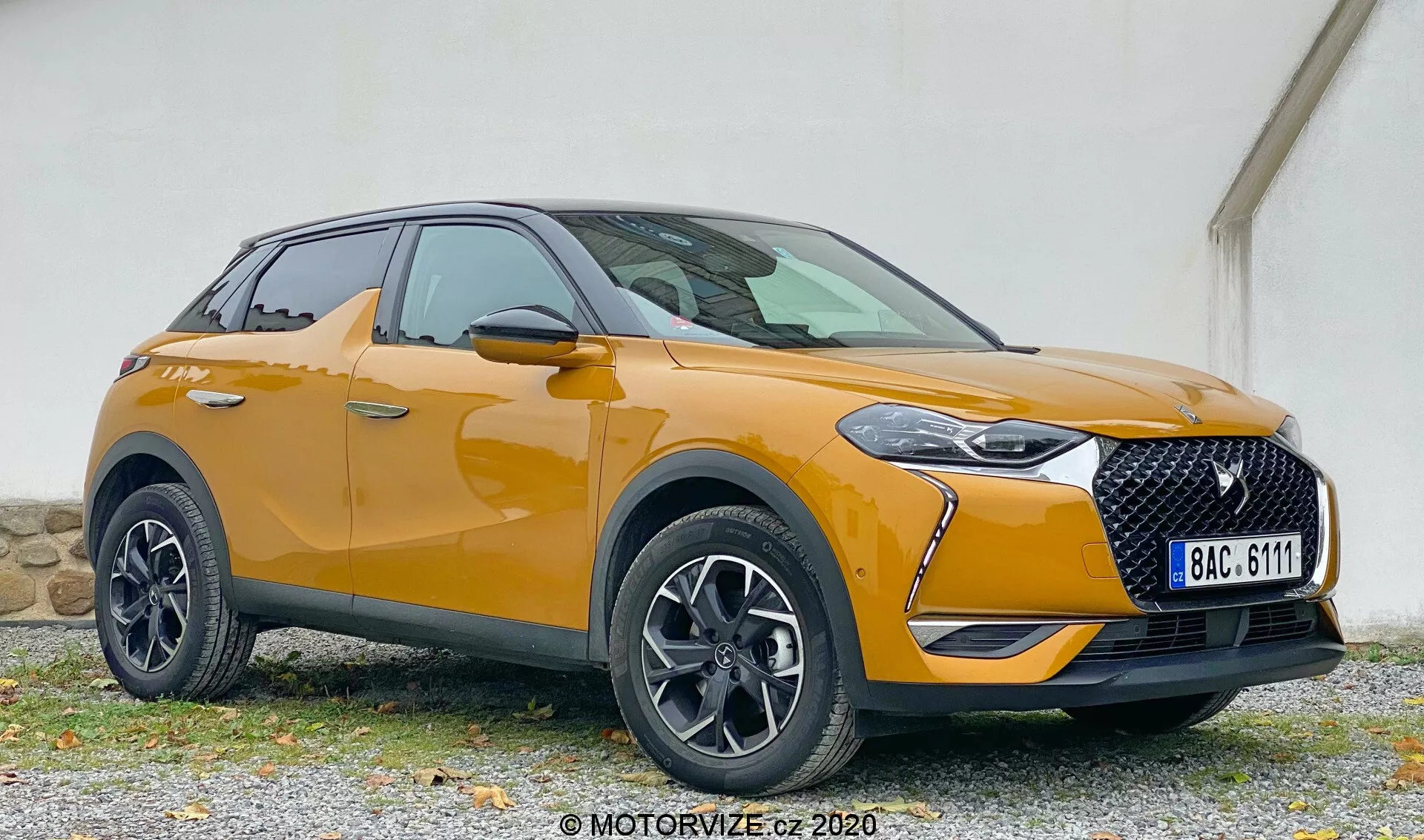 Moderno SUV in vista frontale a tre quarti che mostra la DS Automobiles DS 3 2019 (DS 3 Crossback) con dettagli dell'angolo anteriore sinistro, inclusi faro, griglia, cofano, paraurti anteriore, ruota, parafango anteriore e portiera del conducente, posizionato su una superficie di ghiaia con uno sfondo di parete dai colori tenui.