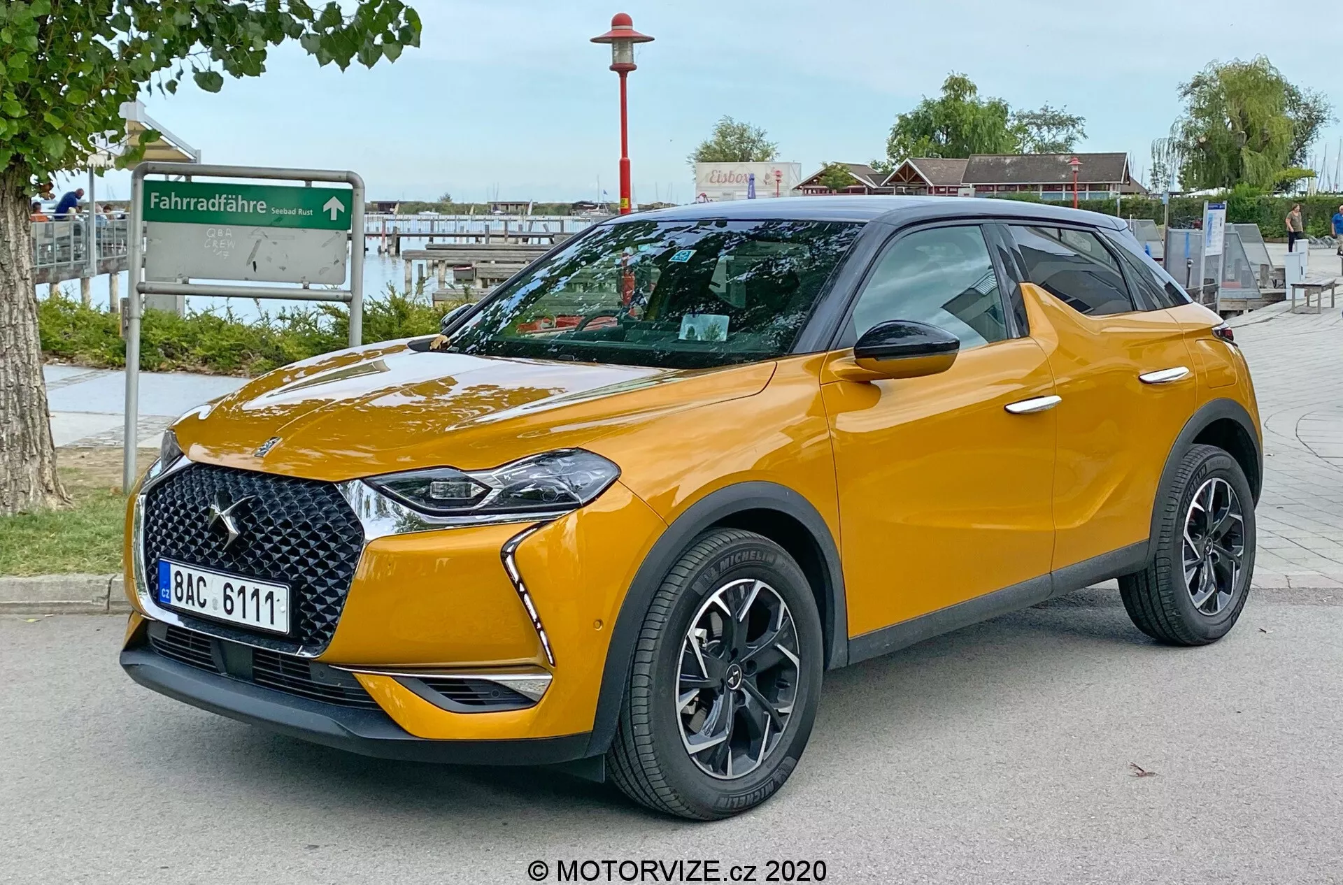Immagine che mostra il moderno SUV DS Automobiles DS 3 Crossback 2019 in una vista anteriore di tre quarti, evidenziando la griglia frontale elegante, i fari LED integrati, il paraurti anteriore, il cofano e il profilo del lato del conducente. Parcheggiato all'aperto su una superficie pavimentata con uno sfondo urbano che include una stazione di condivisione di biciclette. La foto, scattata da un punto di vista basso all'altezza dei fari, accentua l'altezza da terra dell'auto. Le caratteristiche visibili includono ruote in lega di design multi-razze, specchietti con frecce integrate, pneumatici a profilo ribassato, colore del tetto a contrasto con barre sul tetto, finitura della vernice metallizzata, distintivi specifici del trim e vetri posteriori oscurati. La targa è intenzionalmente sfocata per la privacy.