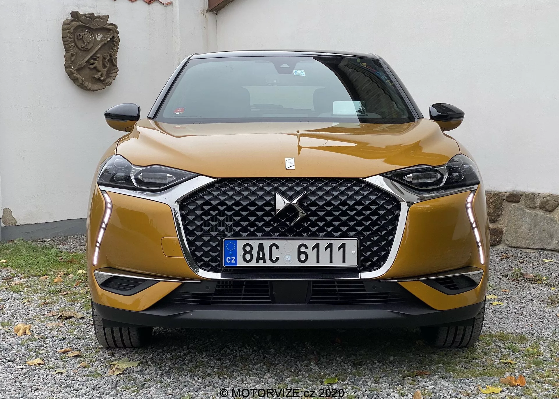 Vista frontale della DS-3 Crossback 2019, che mostra la sua vistosa griglia esagonale con un grande logo centrale affiancata da eleganti unità di fari angolari con probabile tecnologia LED o HID. Il paraurti anteriore sportivo con finiture a contrasto e le linee scolpite del cofano aggiungono dinamismo estetico. L'auto leggermente spostata a destra è fotografata all'altezza della griglia, catturando il design moderno e la tecnologia.