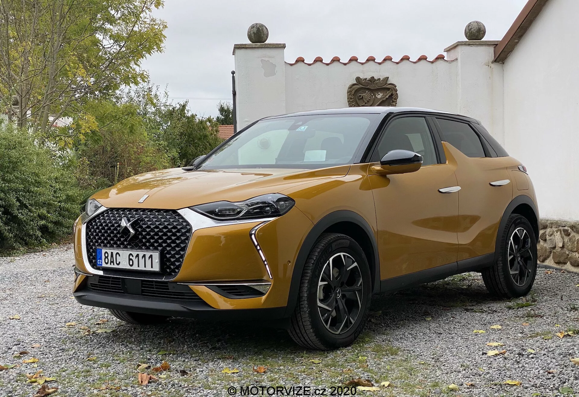 Vista frontale di tre quarti del DS-3 Crossback 2019, un moderno crossover SUV compatto. L'angolo leggermente basso enfatizza l'altezza e l'aspetto robusto, sotto un'illuminazione soffusa e diffusa. Le caratteristiche salienti includono luci diurne a LED distintive intorno al gruppo ottico principale, una grande griglia a motivo con badge prominente, sezioni dei fendinebbia oscurate nel paraurti e una presa d'aria notevole sotto la griglia. Il cofano presenta contorni sottili, le ruote sono in lega nera multirazza, indicative di un livello di finitura superiore, e gli specchietti laterali in tinta carrozzeria, forse con indicatori di direzione integrati. Le linee del corpo sono fluide e le leggere bombature sui passaruota conferiscono robustezza e sportività. La verniciatura metallica suggerisce un livello premium. Il veicolo potrebbe disporre di tecnologia di illuminazione avanzata, sensori di parcheggio e possibilmente sistemi di telecamere o radar integrati, anche se non chiaramente visibili. Situato su una superficie di ghiaia con vegetazione e una struttura edile sullo sfondo, la targa in stile europeo è visibile ma alterata.