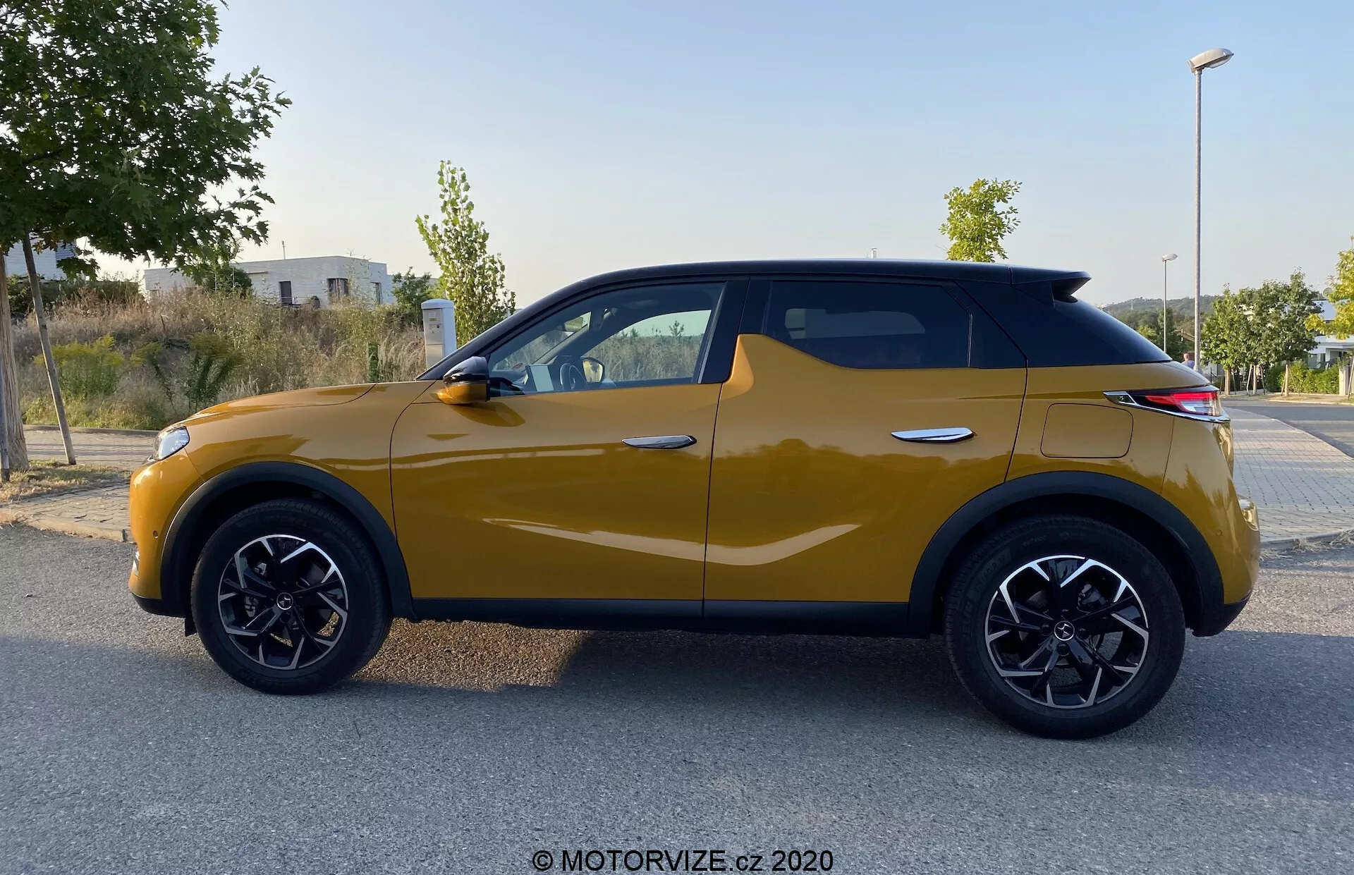 Vista di profilo laterale di una DS Automobiles DS 3 Crossback del 2019 in giallo senape. Il moderno SUV compatto presenta distintivi passaruota con finiture nere, cerchi in lega neri a più razze e una linea dinamica della carrozzeria dal passaruota anteriore ai fanali posteriori. Gli specchietti laterali integrano indicatori di direzione e le finestre sono contornate di nero, evidenziando un design coerente, mentre il tetto che degrada dona un tocco contemporaneo. L'immagine, scattata con luce diffusa, non presenta ombre o riflessi, concentrando l'attenzione sugli elementi del design del veicolo.