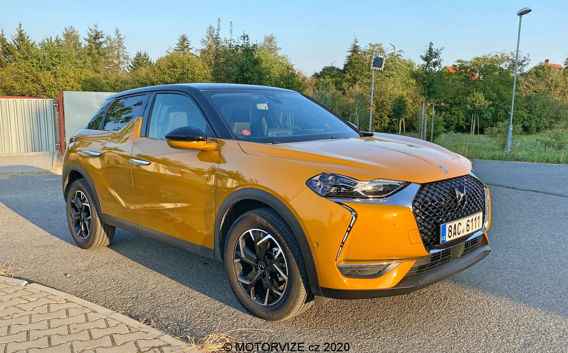 Un SUV compatto e moderno DS Automobiles DS-3 Crossback del 2019, mostrato parcheggiato ad angolo con attenzione al profilo anteriore e laterale. L'immagine, catturata da una prospettiva più bassa vicino ai mozzi delle ruote, mette in evidenza il corpo metallizzato arancione brillante del veicolo e il tetto nero. La parte anteriore presenta una grande griglia nera a motivo diamantato, fari affilati con accenti LED e fari fendinebbia in cornici nere. L'auto ha specchietti laterali neri, finiture cromate per le finestre e ruote nere con accenti argentati. Notevole la posizione leggermente elevata, le linee sportive della carrozzeria e la piega verso il basso attraverso le maniglie delle porte.