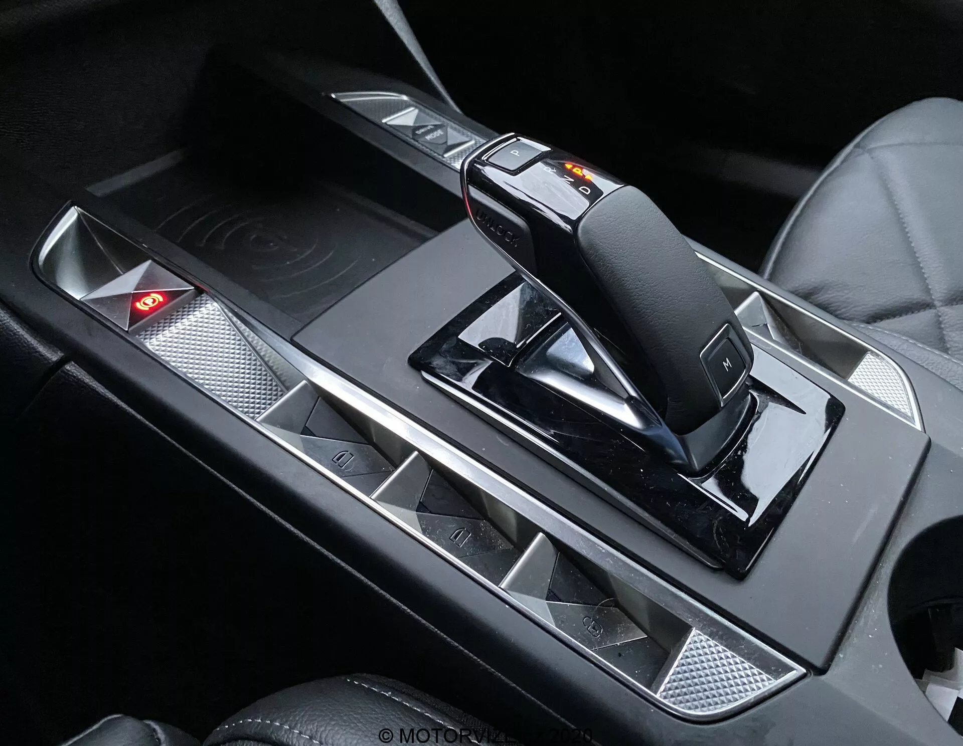 Primo piano della console centrale del DS Automobiles DS-3 Crossback 2019, con particolare attenzione alla moderna e ergonomica leva del cambio con interruttore per la modalità manuale e meccanismo di rilascio. La console è decorata con accenti metallici a contrasto con le superfici nere, portabicchieri con finitura opaca e un pannello di controllo con spia luminosa rossa accesa.