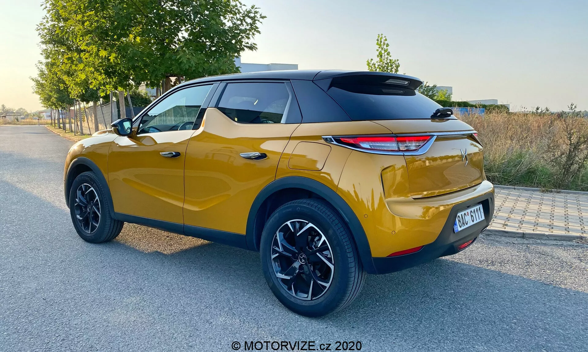 Vista posteriore tre quarti di un moderno crossover SUV alla luce del giorno naturale, con colore oro metallico vibrante e tetto nero sportivo, luci posteriori a LED moderne, grandi cerchi in lega neri, altezza da terra elevata, sensori di parcheggio posteriori visibili e design bicolore, indicativo di un DS Automobiles DS 3 Crossback 2019.