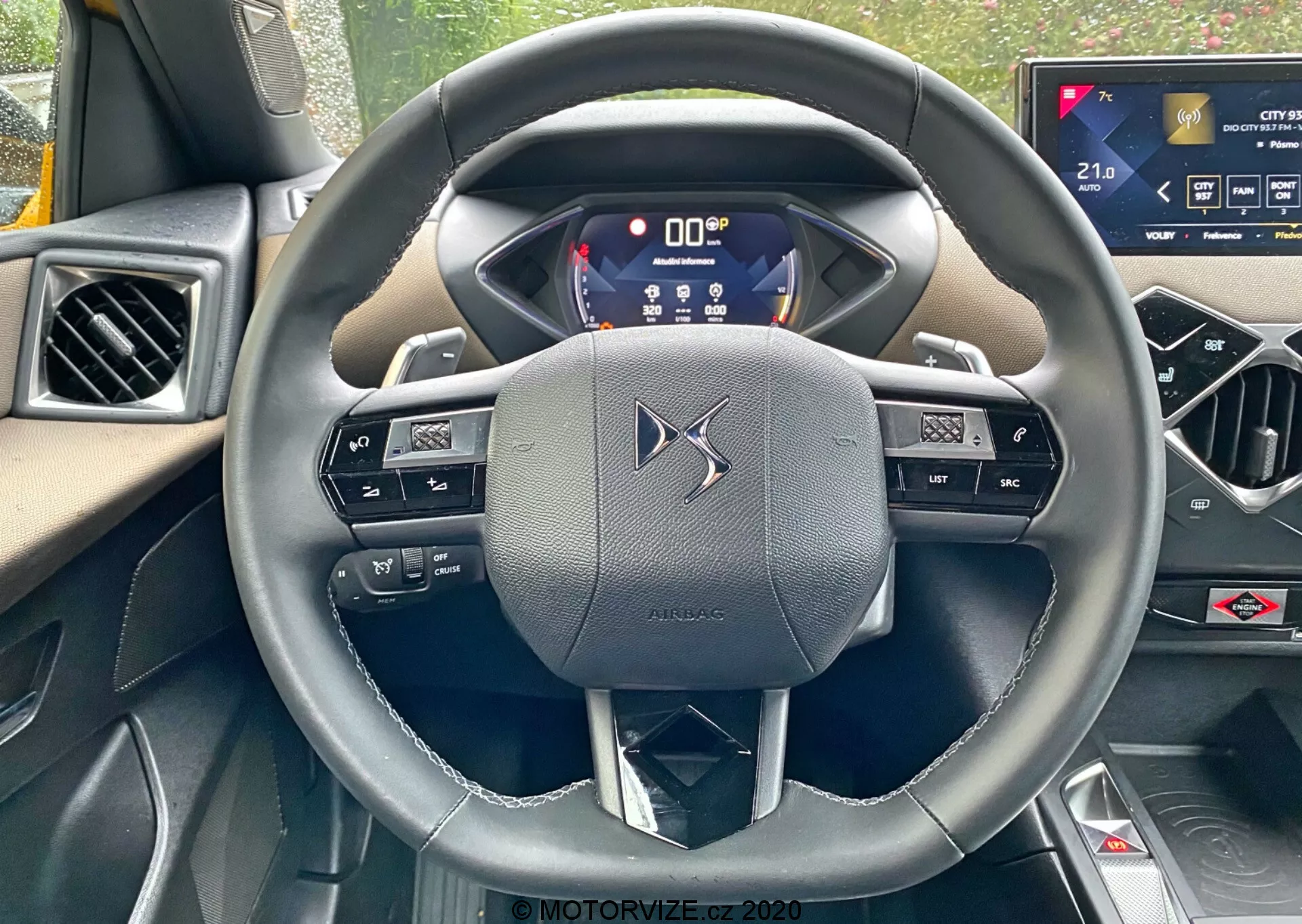 Prospettiva in prima persona dal sedile del conducente di una DS-Automobiles DS-3 2019 (DS-3 Crossback), focalizzata su un volante a tre razze rivestito in pelle con comandi integrati e paddle shifters. Il quadro strumenti digitale mostra informazioni critiche, evidenziando la tecnologia avanzata del veicolo. Un sistema di infotainment touchscreen sulla console centrale suggerisce le caratteristiche di alta comodità della vettura.