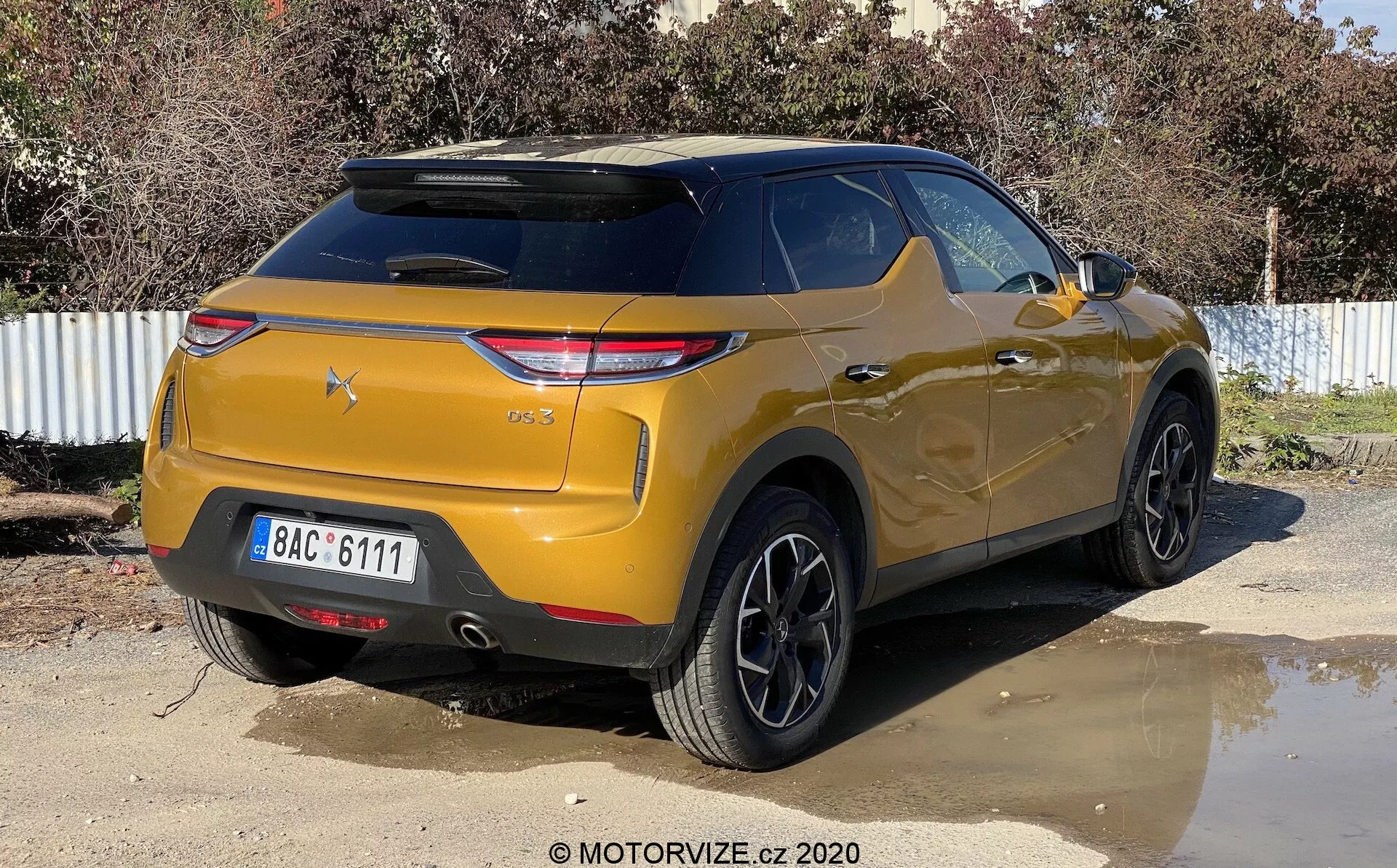 Una vista posteriore di tre quarti di un SUV DS-3 Crossback di colore oro metallizzato parcheggiato all'aperto in una giornata soleggiata. L'immagine cattura il veicolo da un angolo leggermente destro, evidenziando il lato posteriore destro e l'area del bagagliaio. Le caratteristiche principali includono gruppi di luci posteriori angolari moderne, un parabrezza posteriore inclinato con uno spoiler integrato, un paraurti prominente con sensori di parcheggio, cerchi in lega neri a più raggi, un'elevata altezza da terra e vetri posteriori oscurati.