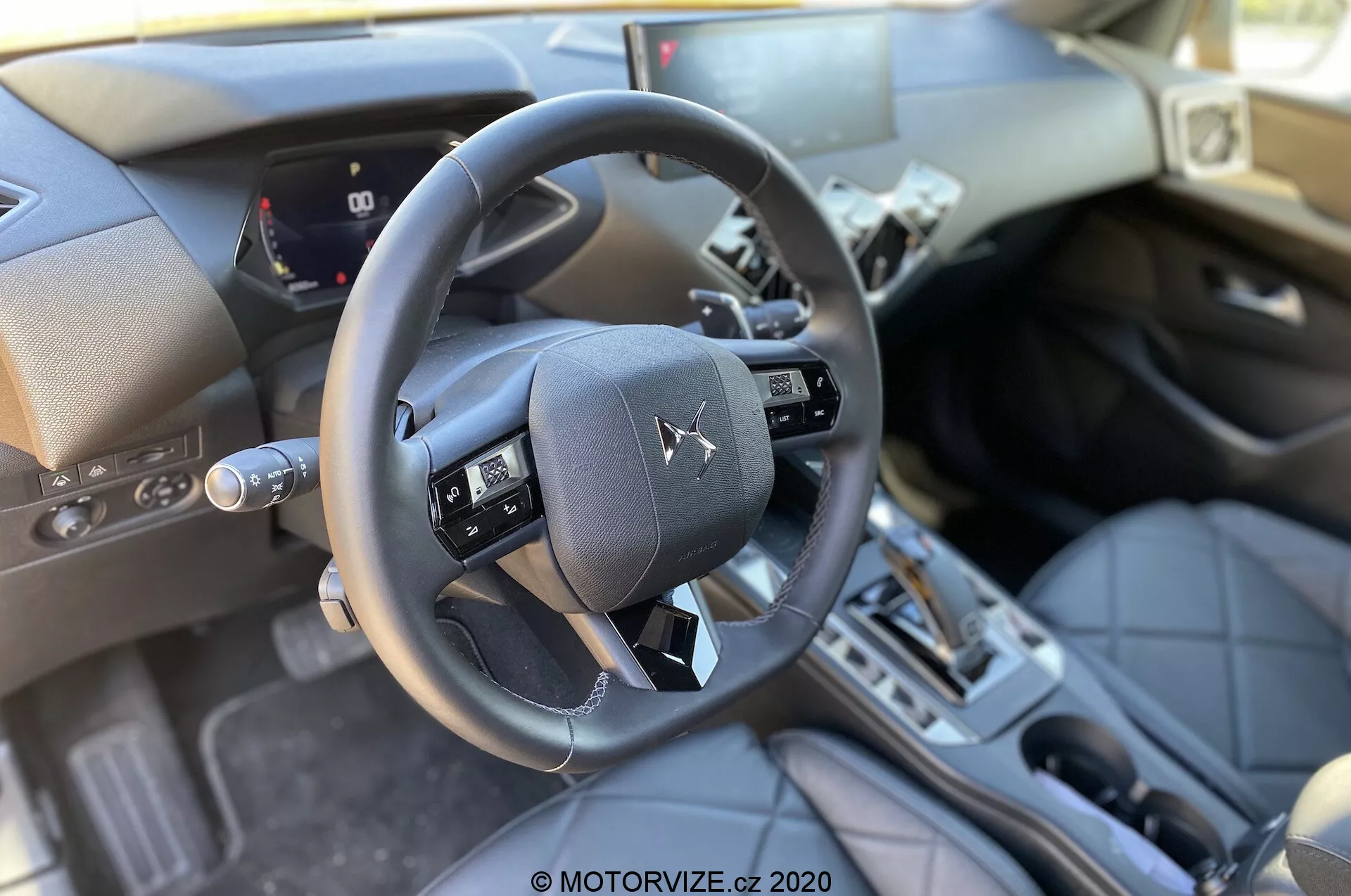 Vista interna della DS Automobiles DS 3 Crossback 2019 dalla prospettiva del sedile passeggero anteriore, evidenziando il volante a tre razze con pulsanti multifunzione, quadro strumenti digitale parziale che mostra la velocità dell'auto e misuratori, console centrale con leva del cambio automatico, materiali premium come superfici in pelle e accenti metallici, slot di accensione tradizionale, pedali e illuminazione naturale e luminosa.
