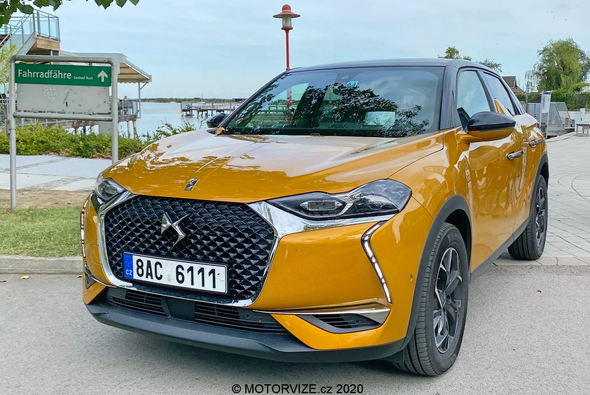 L'immagine mostra un DS Automobiles DS 3 Crossback 2019, un moderno SUV crossover compatto, parcheggiato su una strada asfaltata. Fotografato da un angolo a tre quarti frontale basso, l'immagine mette in evidenza la fascia anteriore e il lato passeggero del veicolo. Le caratteristiche principali includono un emblema del marchio prominente sulla griglia a motivo diamante, fari a LED eleganti con luci diurne stilizzate, fari fendinebbia integrati nel paraurti anteriore, specchietti laterali in tinta con gli indicatori di direzione e ruote in lega multi-razze di colore scuro. L'auto è verniciata in un vivace colore arancione metallizzato con linee della carrozzeria pronunciate che migliorano l'aspetto aerodinamico e distintivi che indicano potenzialmente i livelli di allestimento. L'ambiente suggerisce un'area pubblica vicino a un parco, provata da un segnale di pista ciclabile sullo sfondo.