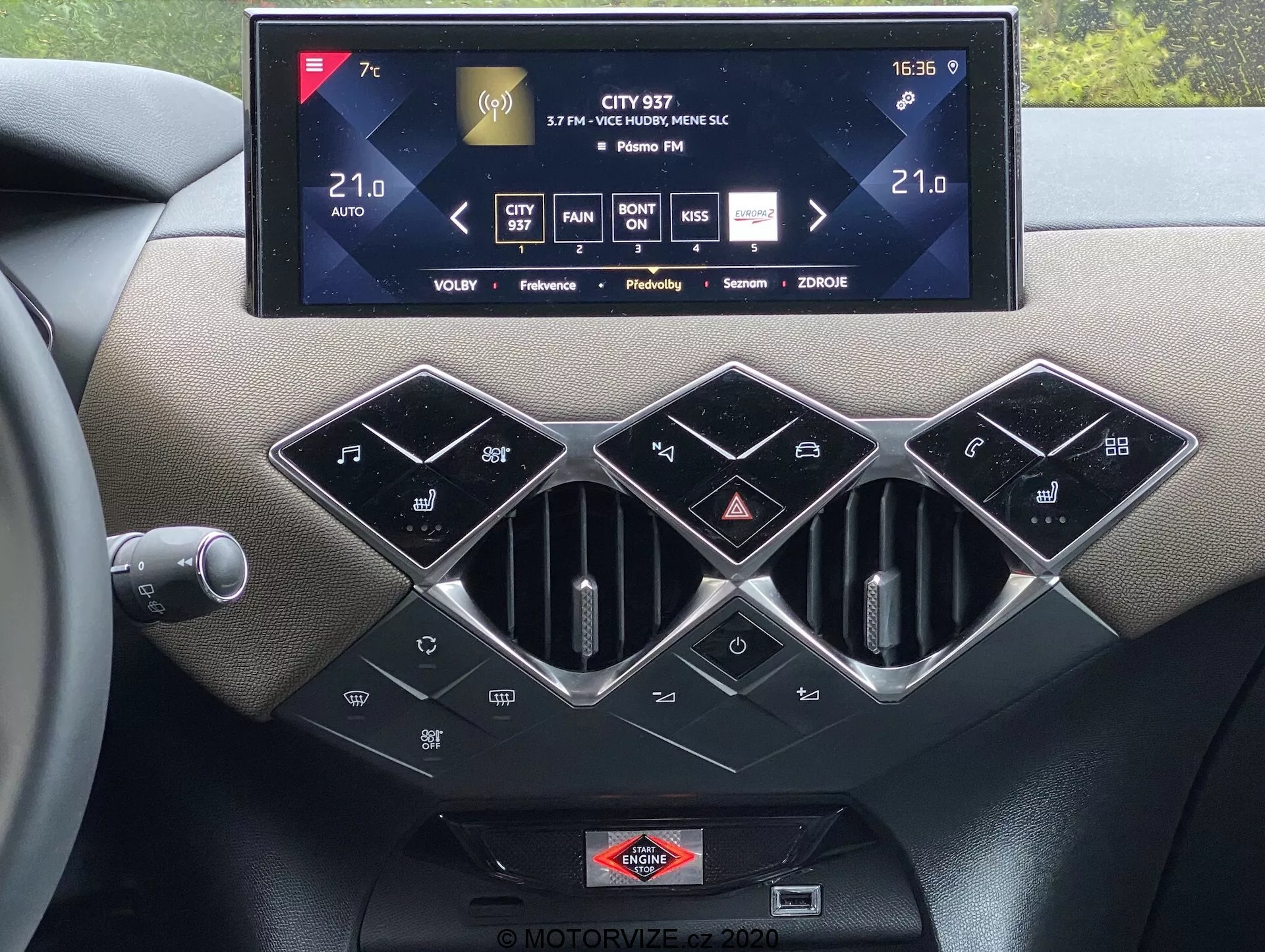 Vista interna della console centrale del ds-automobiles ds-3 2019 che mostra un grande sistema di infotainment touchscreen che mostra il controllo del clima, informazioni sulla radio e impostazioni, con tre prese d'aria esagonali rifinite in argento sotto. C'è una fila di pulsanti di controllo del clima lucidi neri, tra cui SYNC per il controllo del clima multizona, capacità di controllo del clima automatico e una targhetta del marchio posizionata sotto al touch screen, sottolineando materiali tattili di alta qualità e moderna tecnologia automobilistica